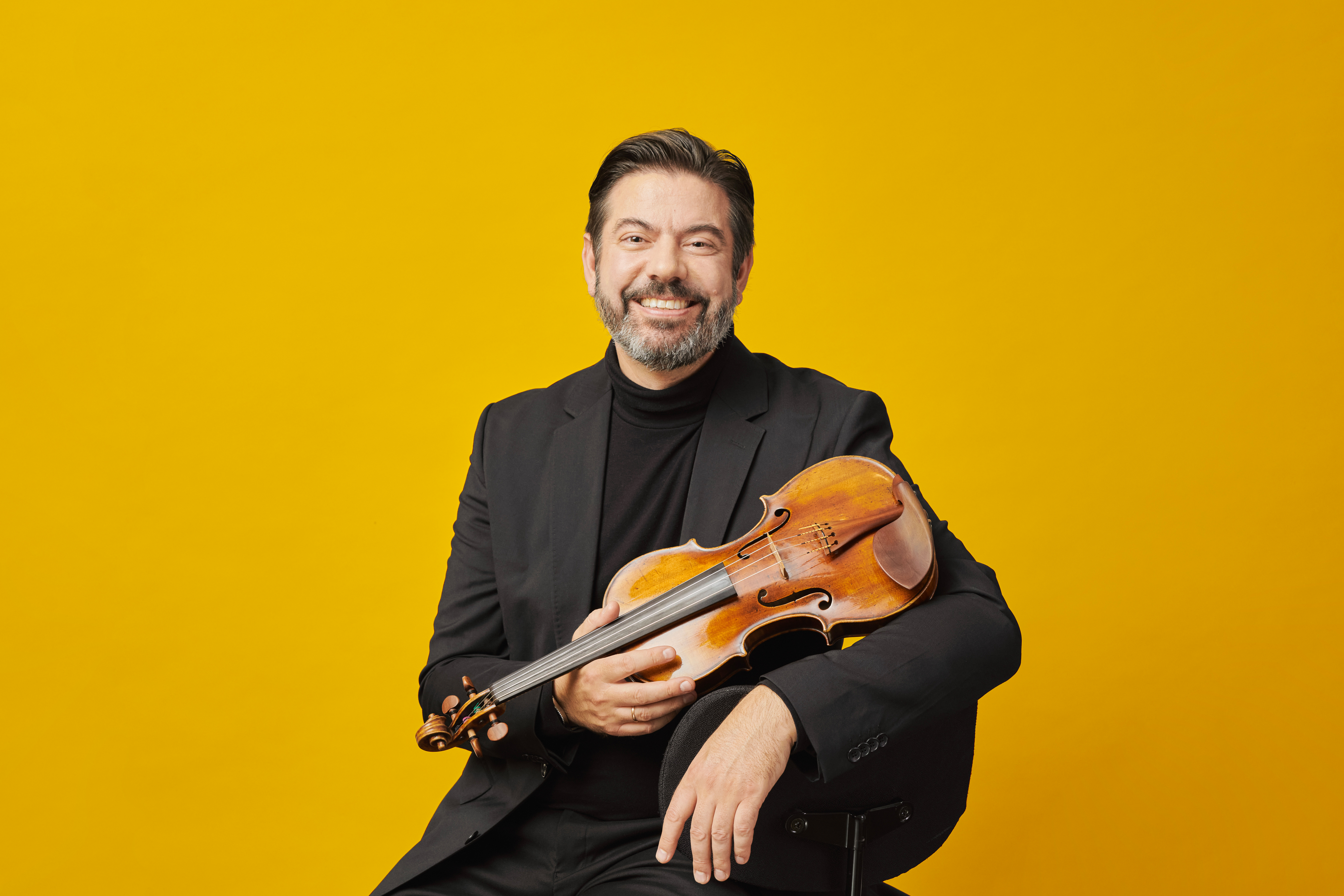 Man with a beard against a yellow background, holding a violin and smiling at the camera.