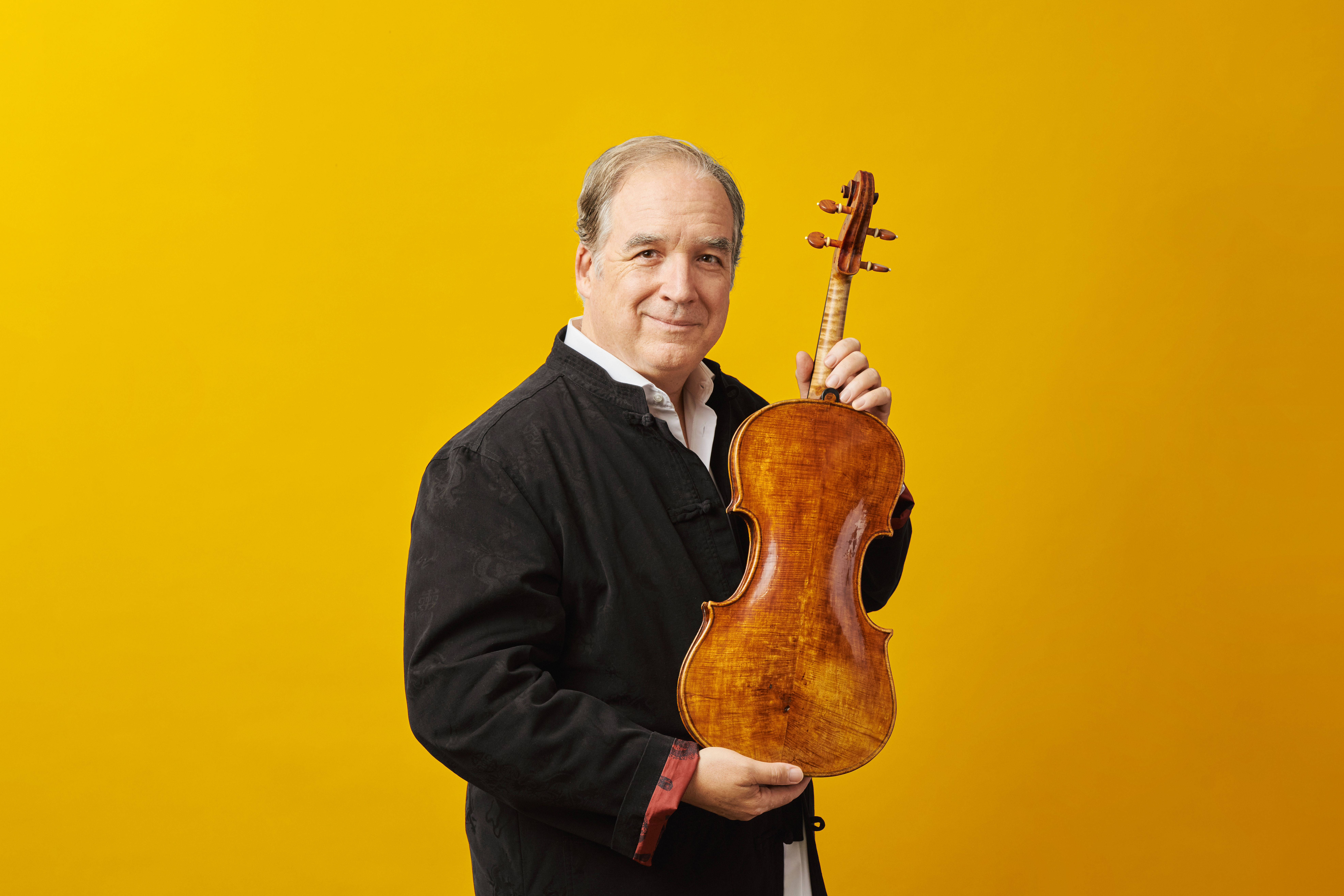 Ein Mann in einer schwarzen Jacke steht vor einem leuchtend gelben Hintergrund, lächelt und hält ein hölzernes Streichinstrument, möglicherweise eine Bratsche, senkrecht am Hals.