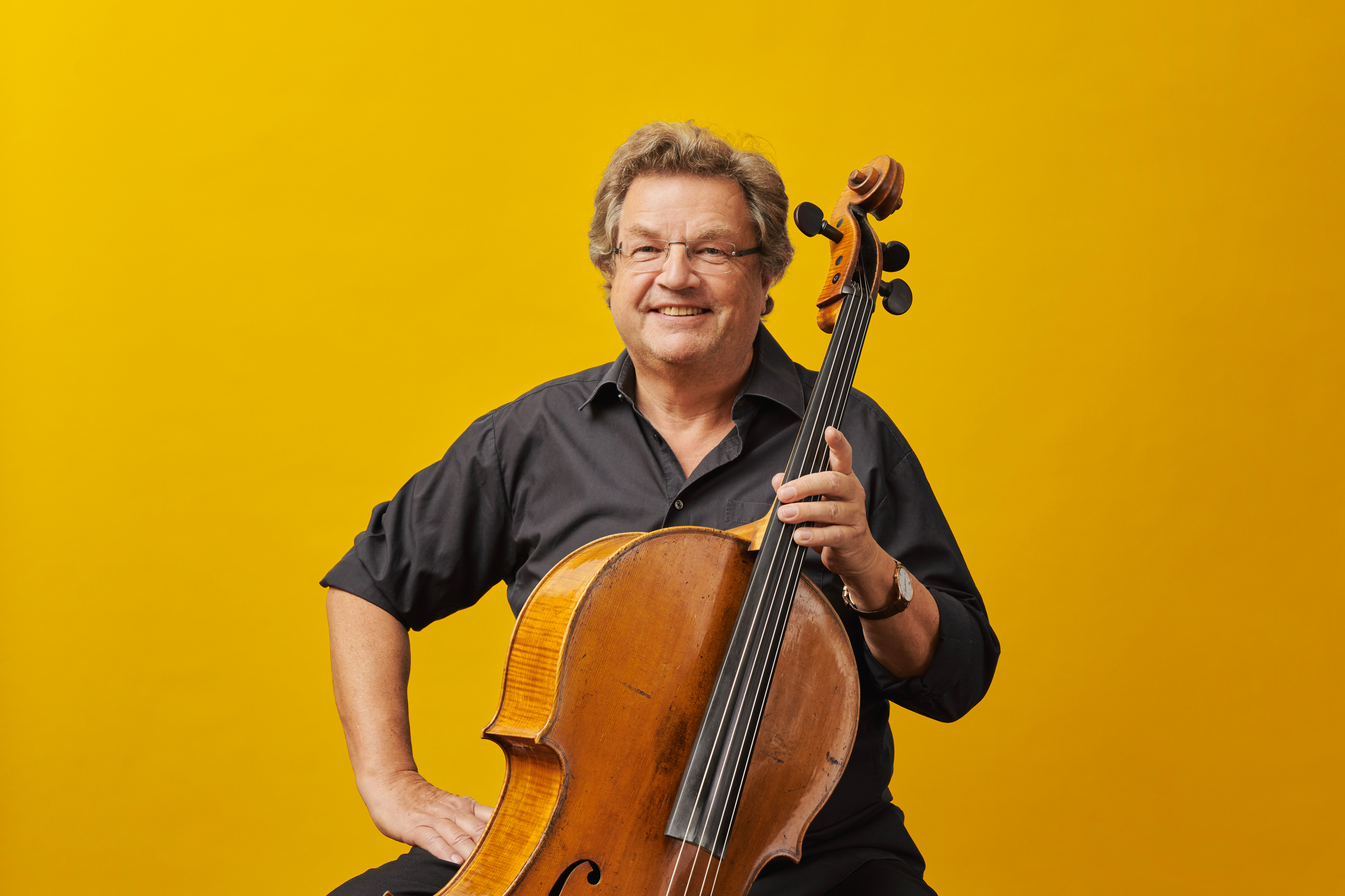 Older cellist wearing glasses against a yellow background, holding his cello and smiling at the camera.