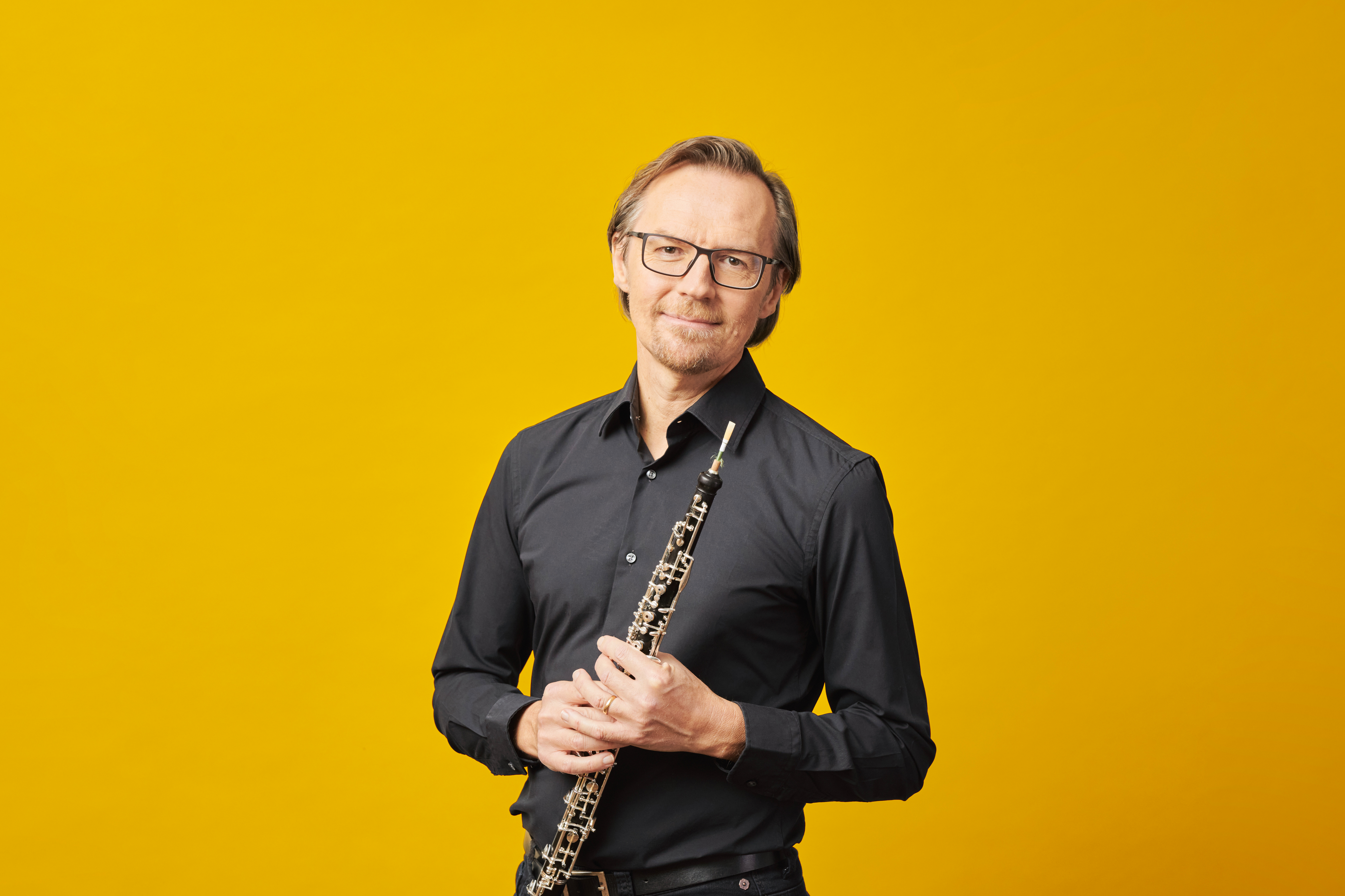 Ein Mann in einem schwarzen Hemd hält eine Oboe vor einem leuchtend gelben Hintergrund.