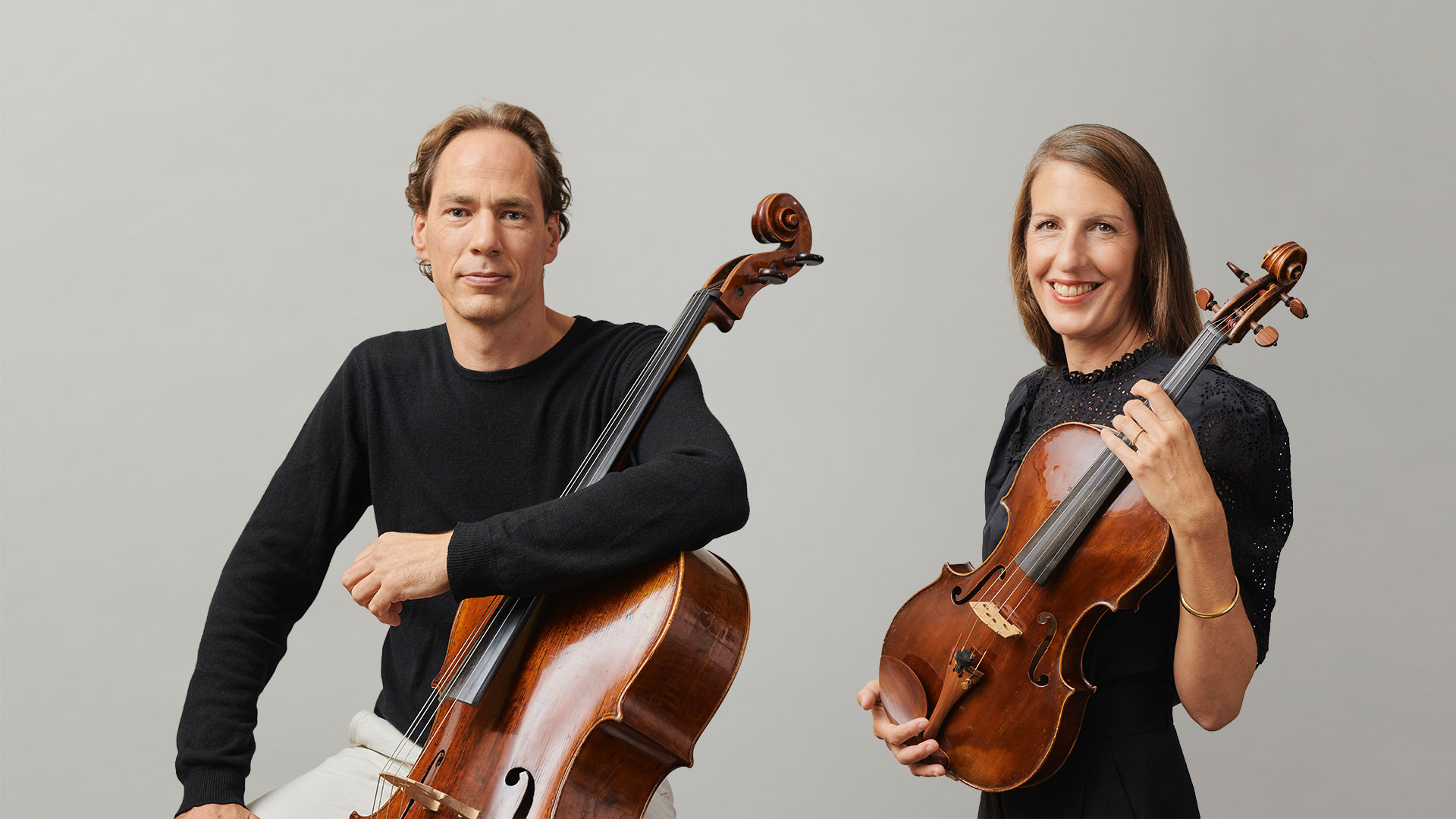 A man and woman pose against a plain background, each holding a string instrument; the man has a cello and the woman has a viola. Both are smiling slightly and wearing dark tops.