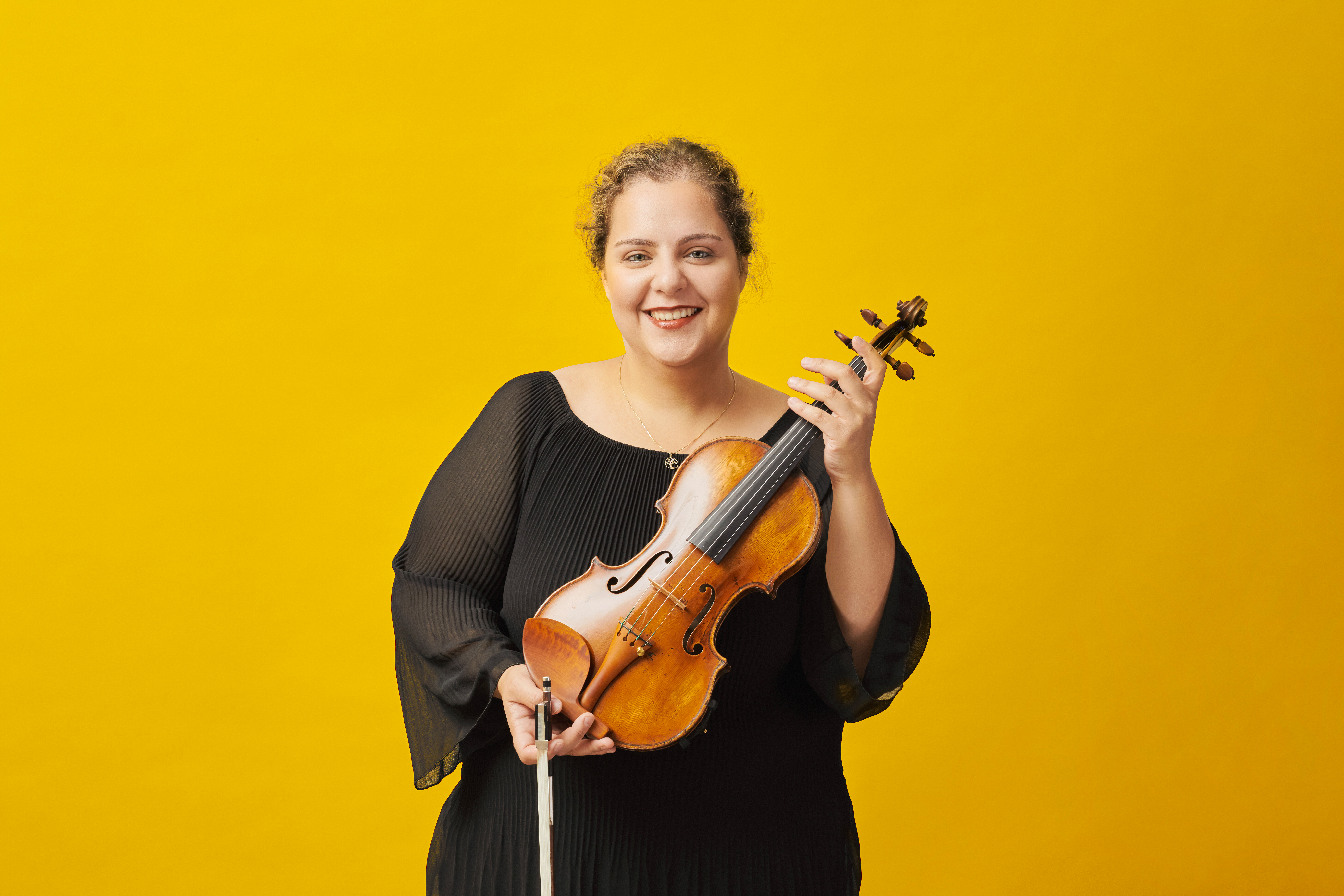 Eine Frau mit lockigem Haar in einem schwarzen Oberteil hält eine Violine vor einem leuchtend gelben Hintergrund.