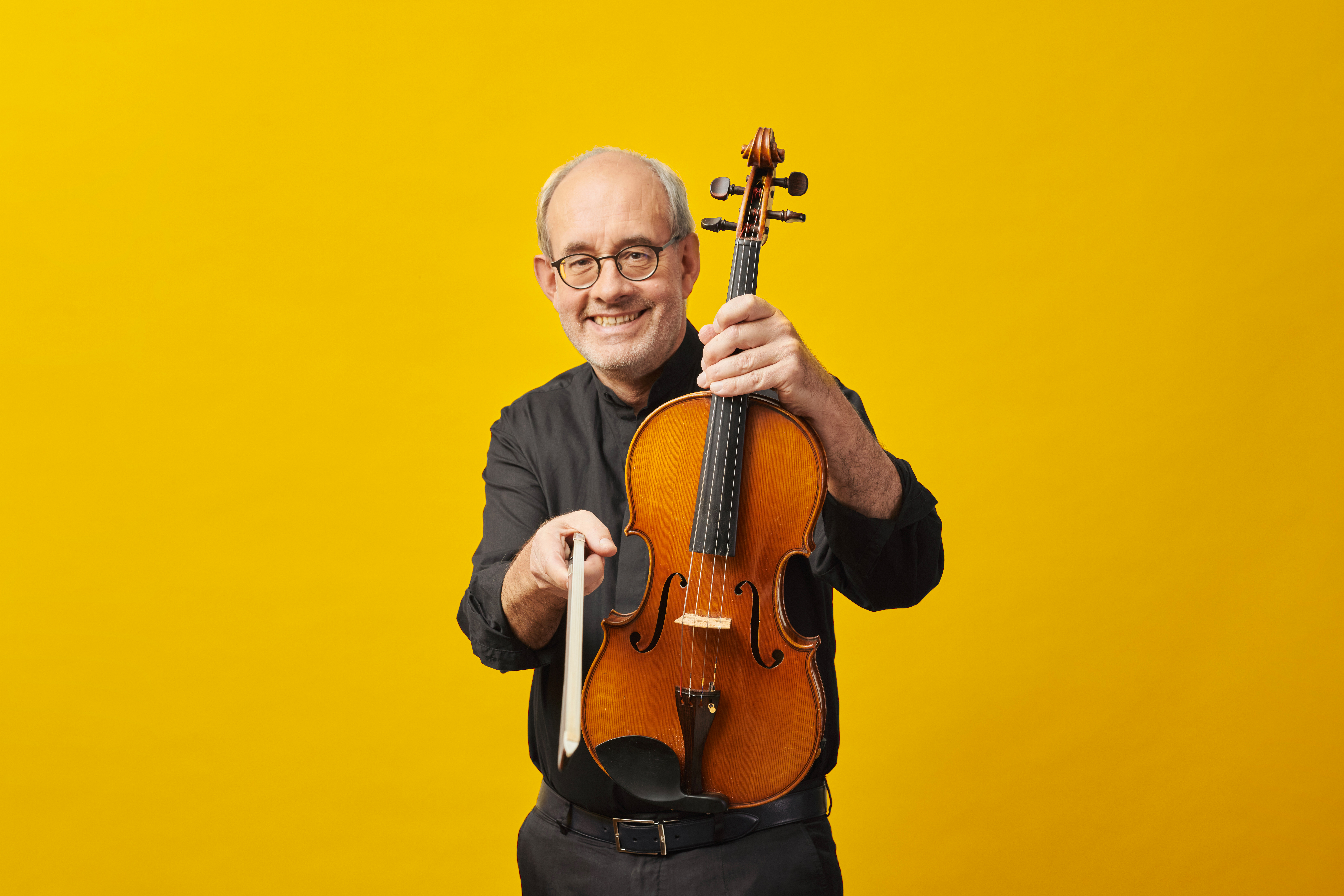 A man wearing a black shirt holds a viola in one hand and points a bow toward the camera with the other, against a bright yellow background.