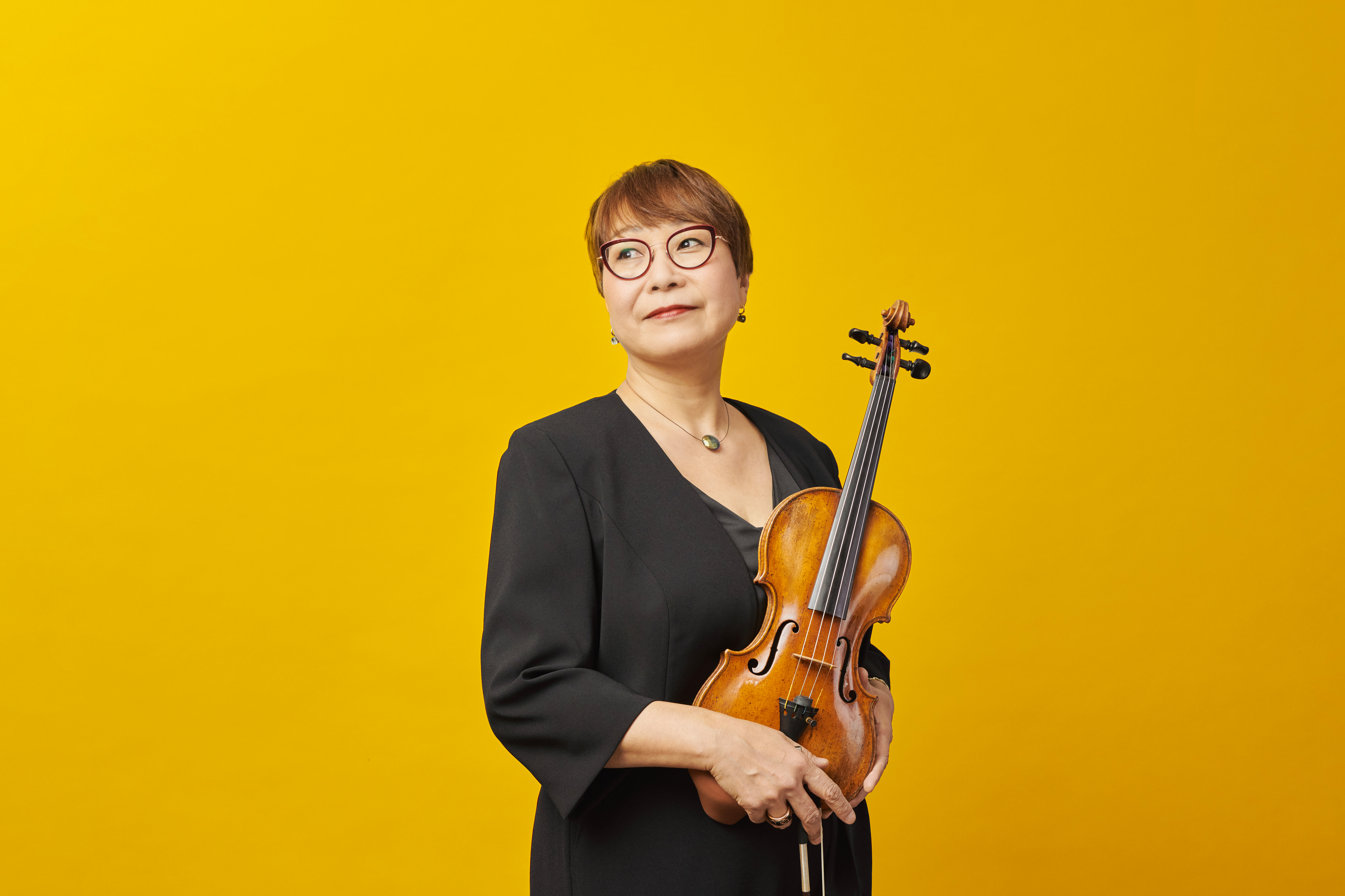 Eine Frau mit kurzen Haaren in einem schwarzen Oberteil hält eine Violine vor einem leuchtend gelben Hintergrund.