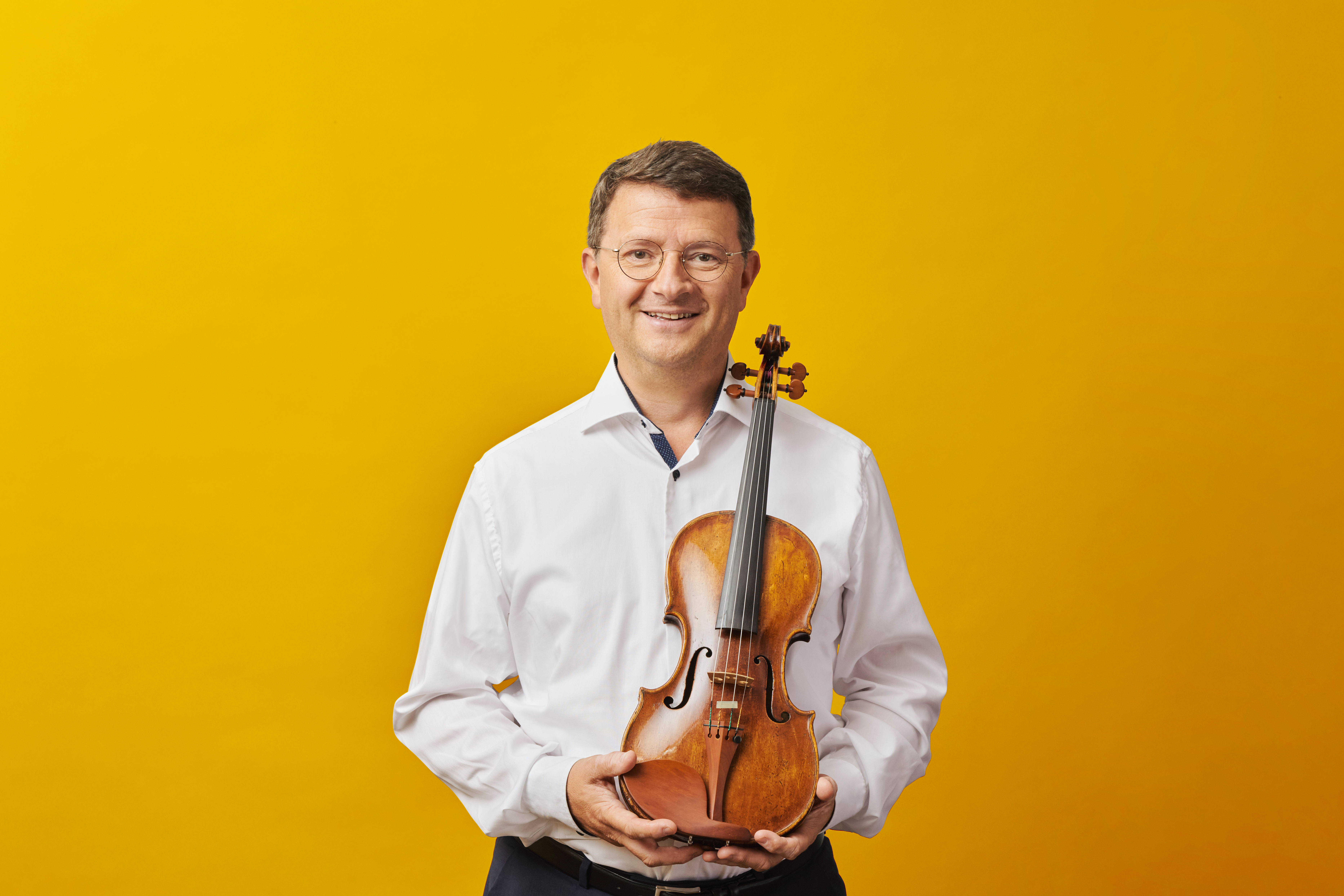 A man wearing a white shirt holds a violin against a bright yellow background.