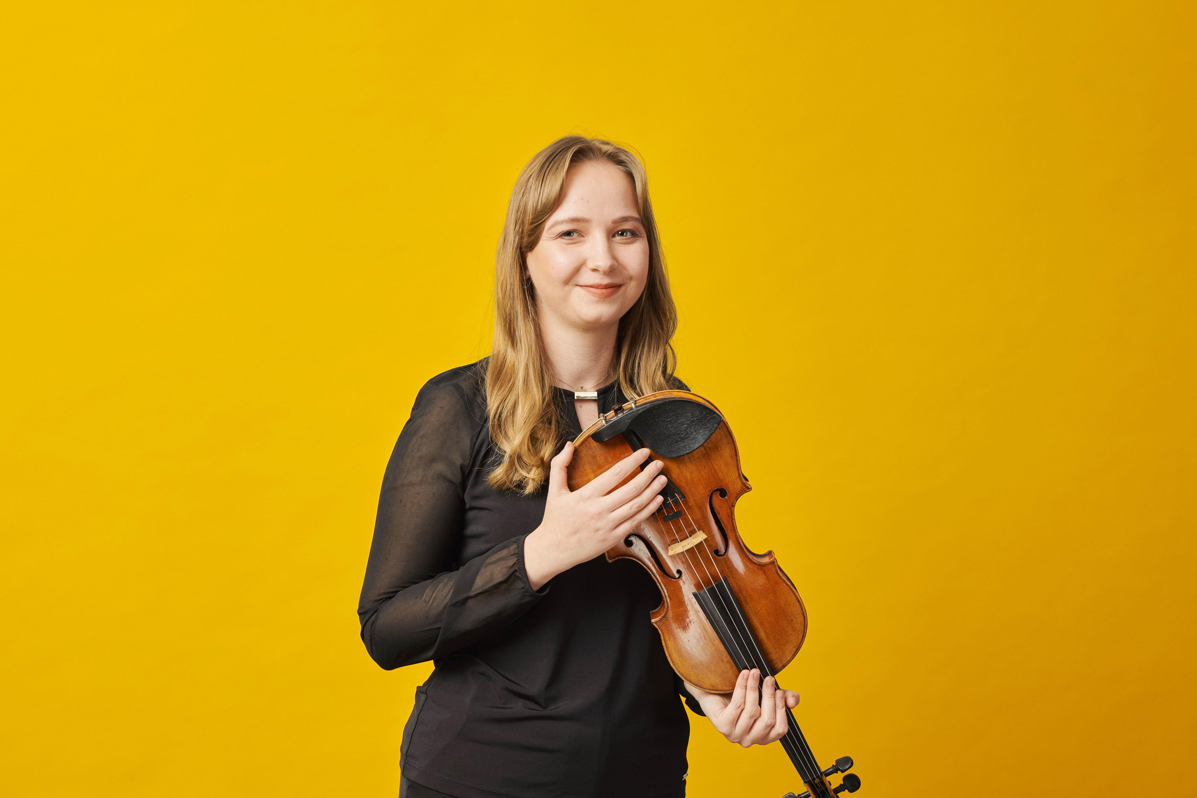 A woman with long blonde hair, wearing a black top, holds a violin and smiles in front of a bright yellow background.