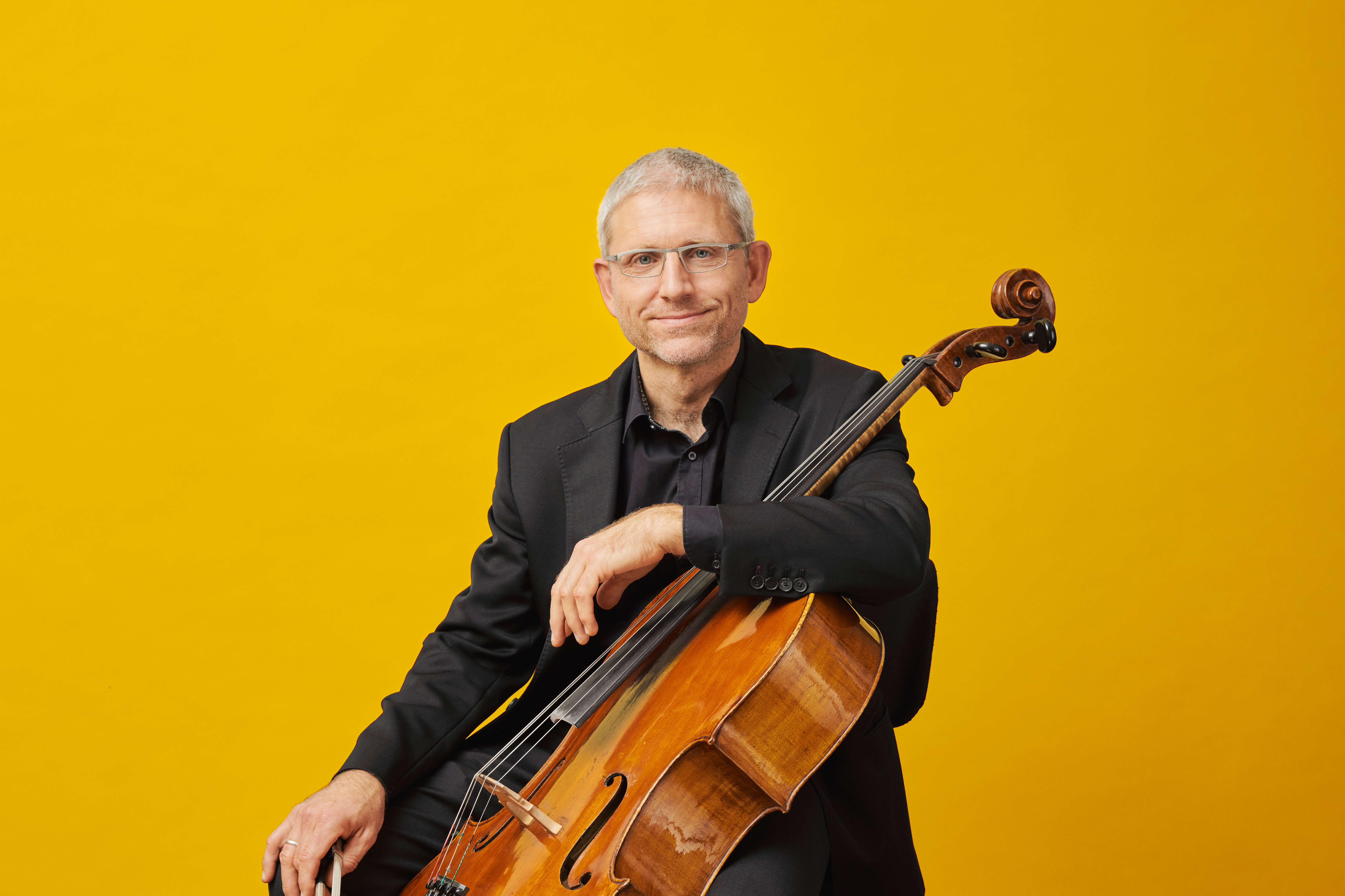 Ein Mann in einem schwarzen Anzug sitzt und hält ein Violoncello vor einem leuchtend gelben Hintergrund.