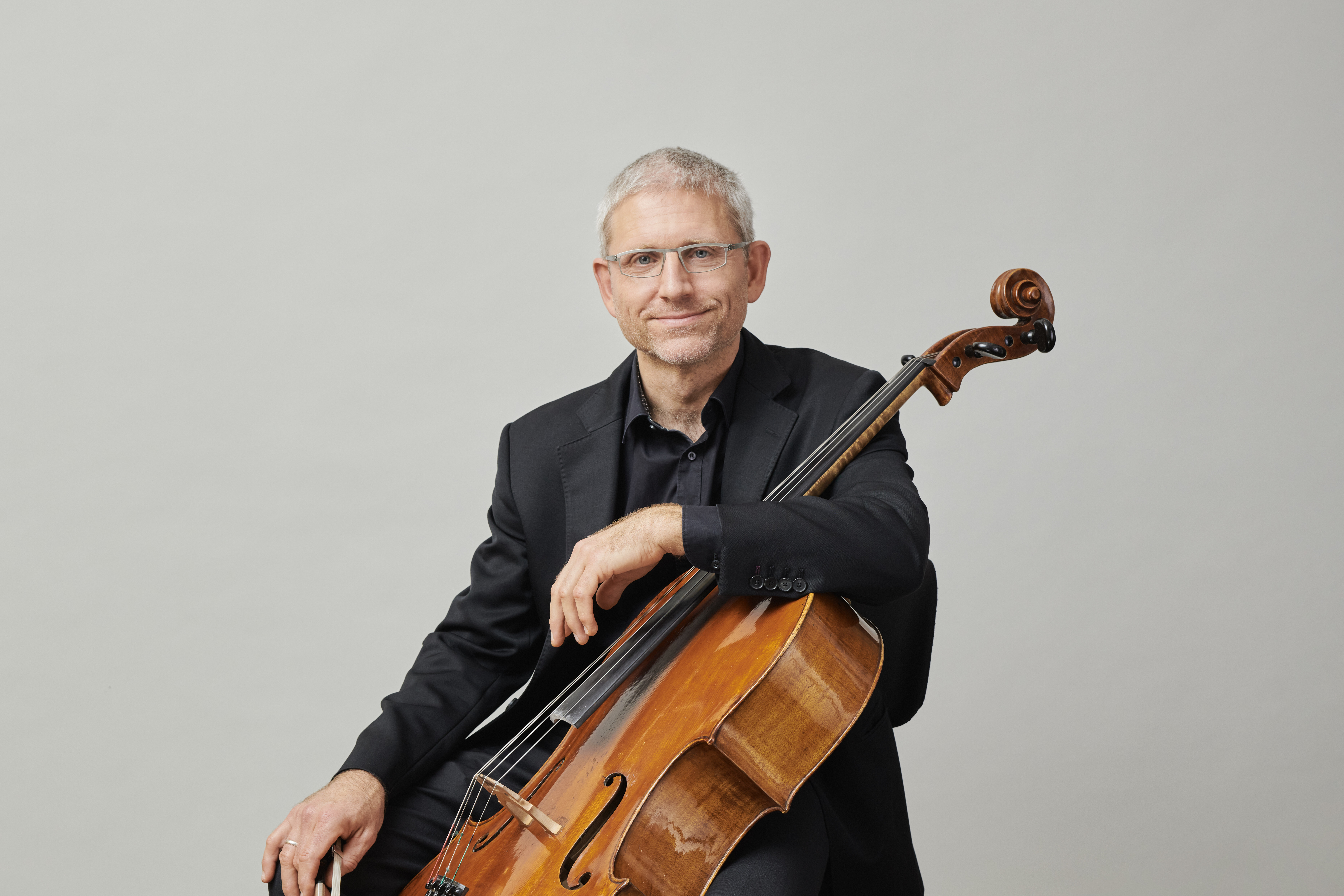 Ein Mann mit kurzen grauen Haaren und Brille, der einen schwarzen Anzug trägt, sitzt und lächelt, während er ein Cello vor einem schlichten hellen Hintergrund hält.