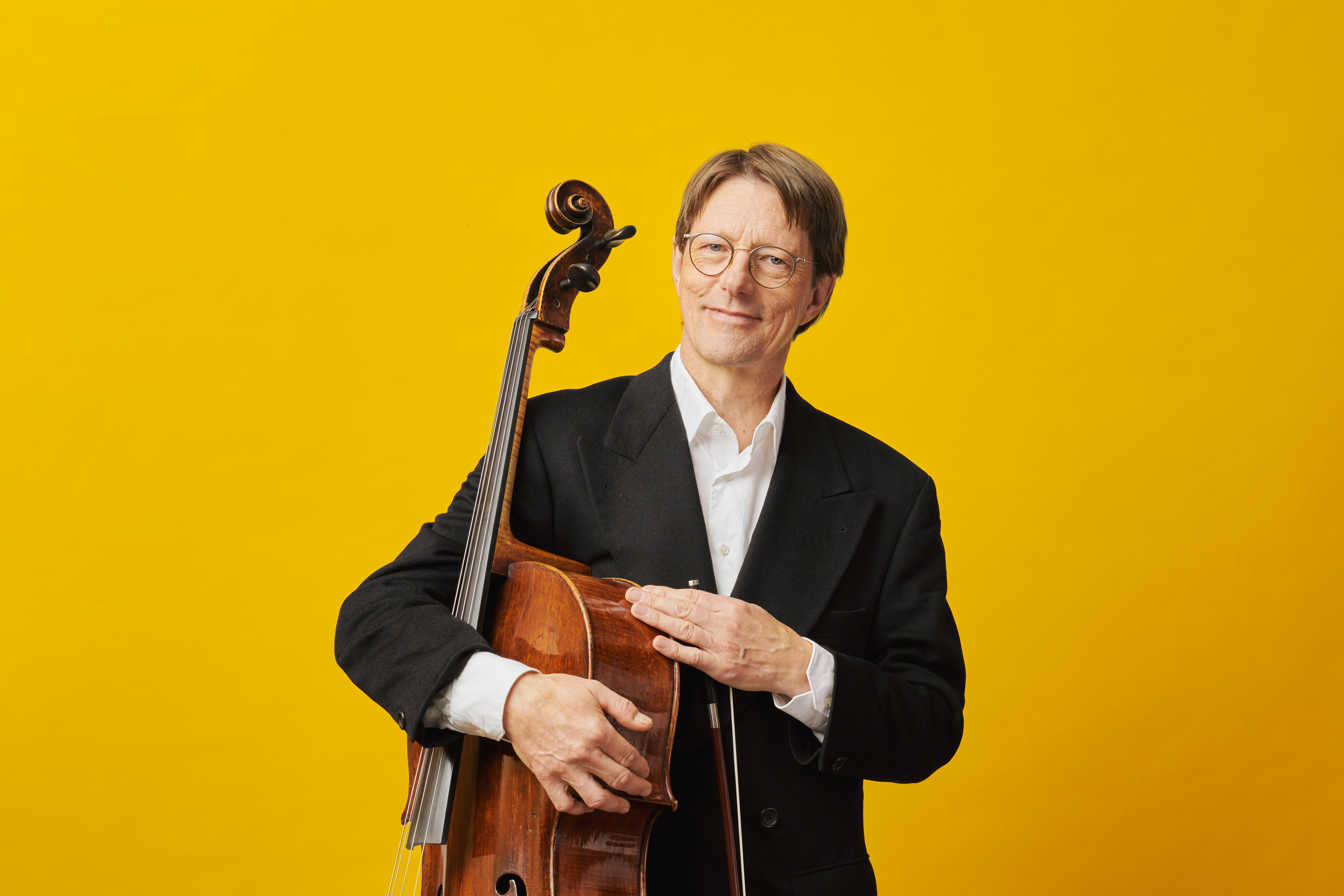A smiling man in glasses and a black jacket holds a cello and bow in front of a bright yellow background.