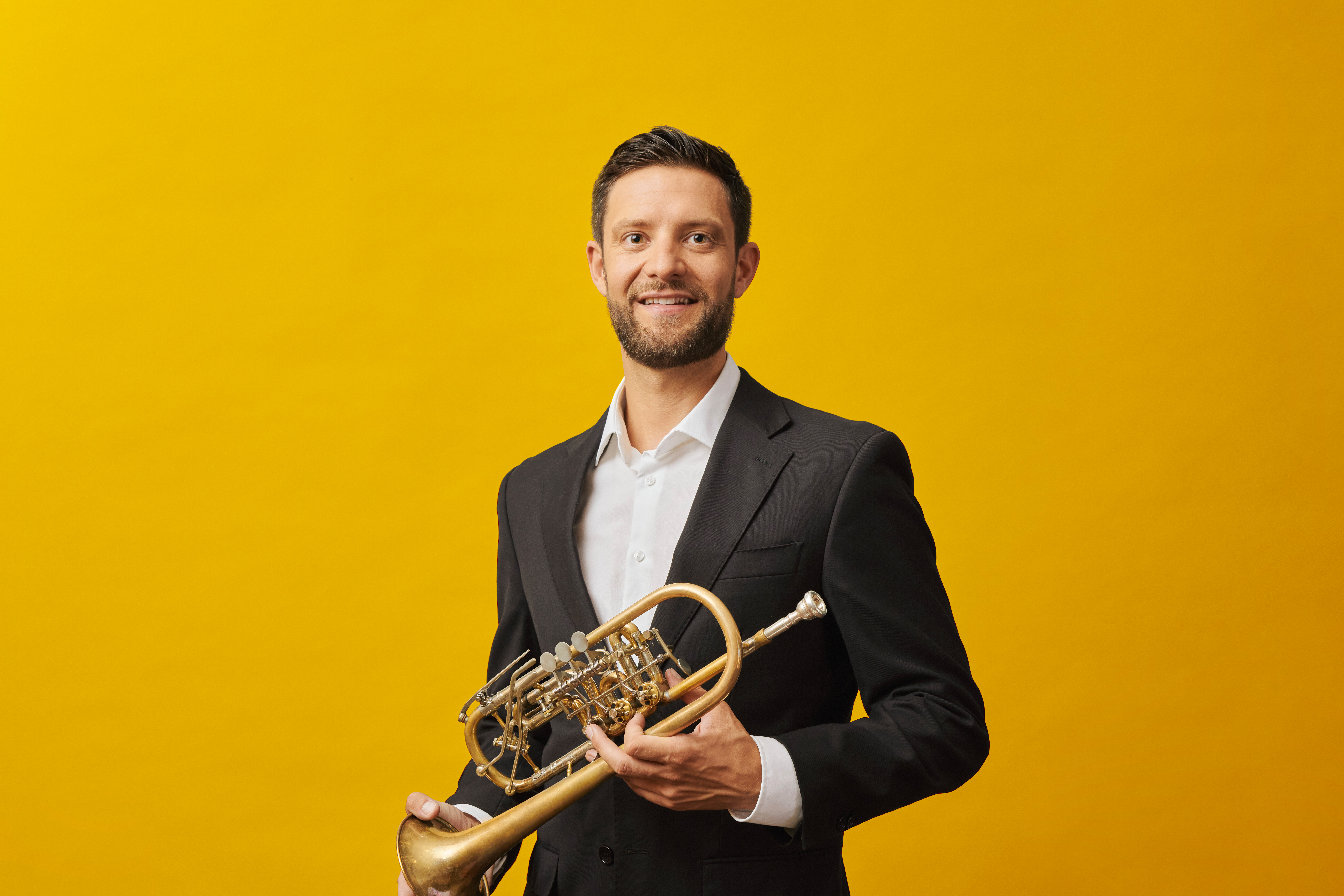 A man in a black suit and white shirt smiles while holding a trumpet against a bright yellow background.