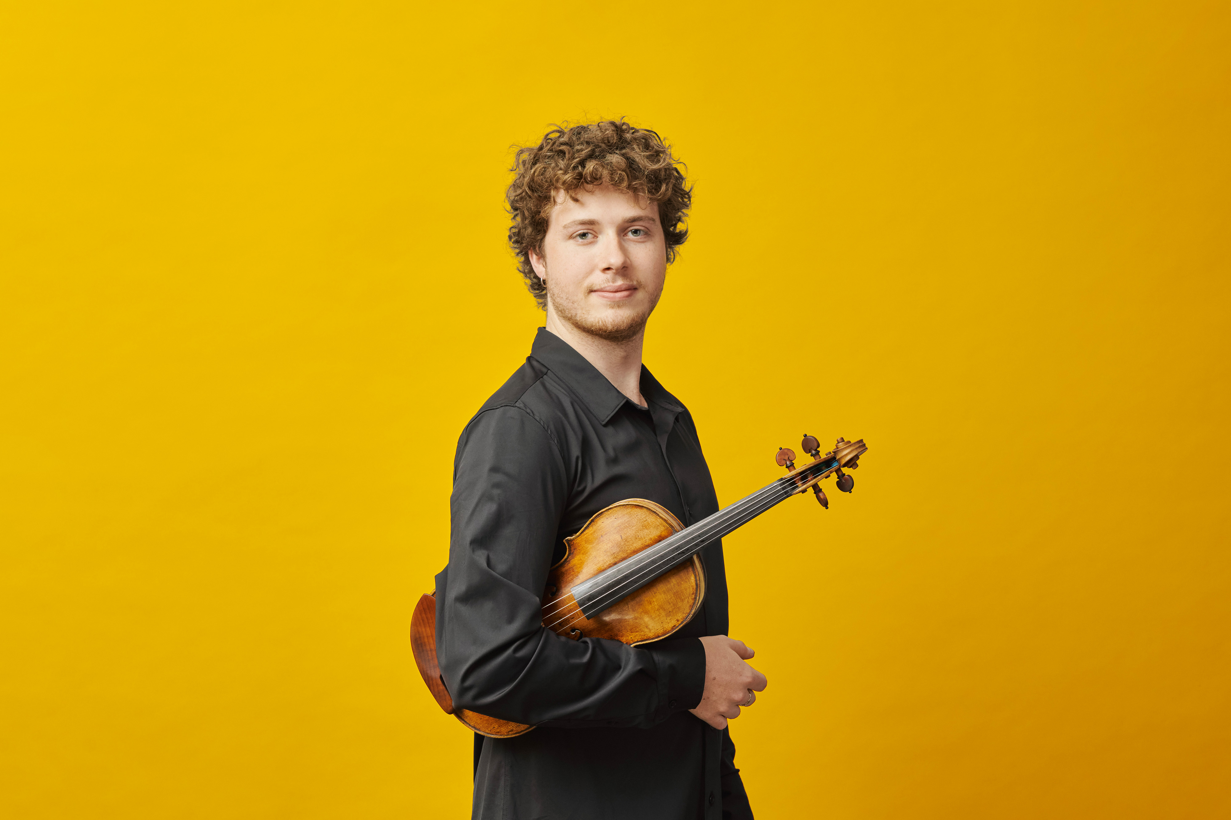 A young man with curly hair wearing a black shirt holds a violin under his arm and stands against a bright yellow background, looking confidently at the camera.