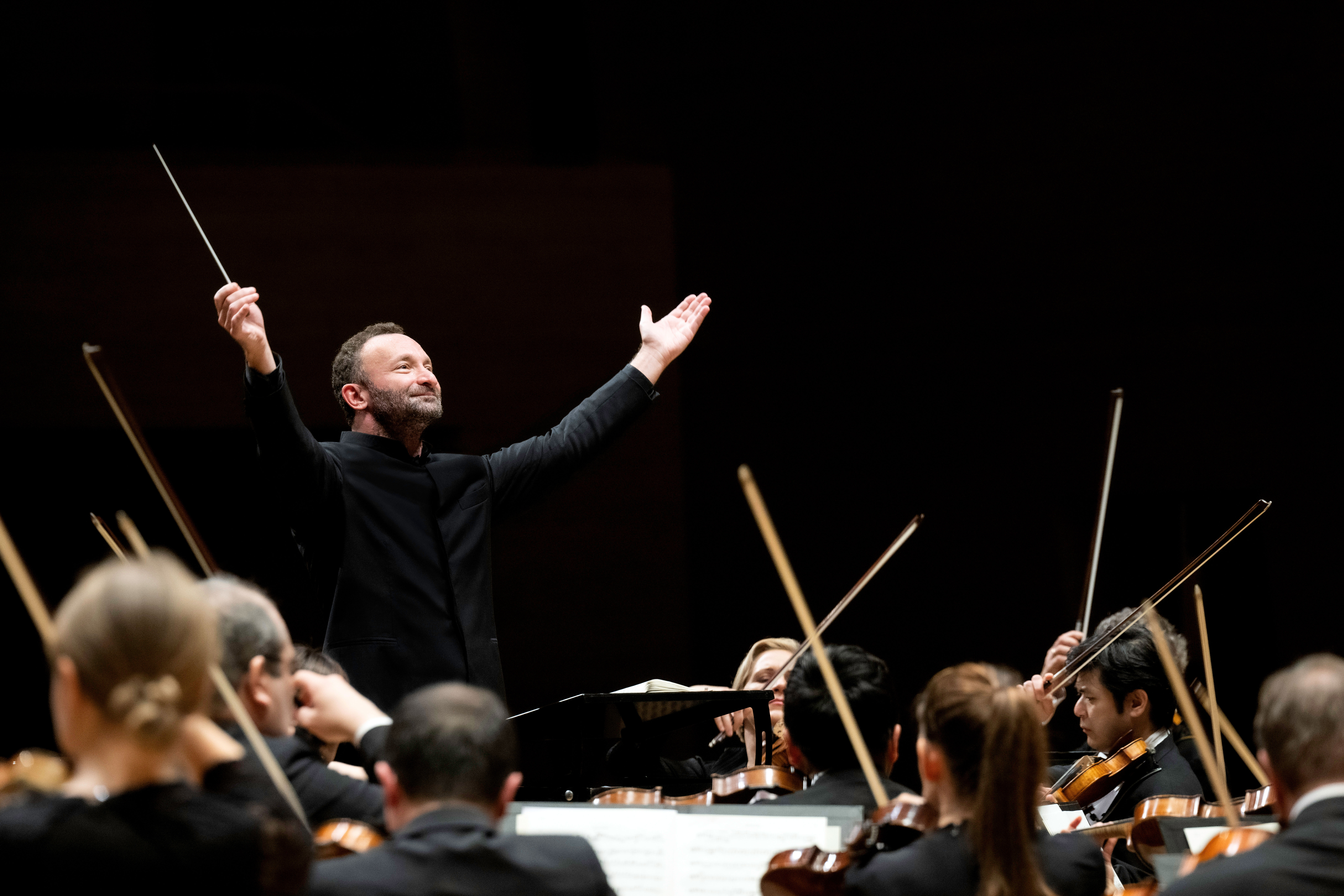 Kirill Petrenko steht vor dem Orchester und hebt beide Arme zu einer großen Geste nach oben. Er trägt einen schwarzen Anzug. Im Vordergrund sind Musiker von hinten zu sehen.