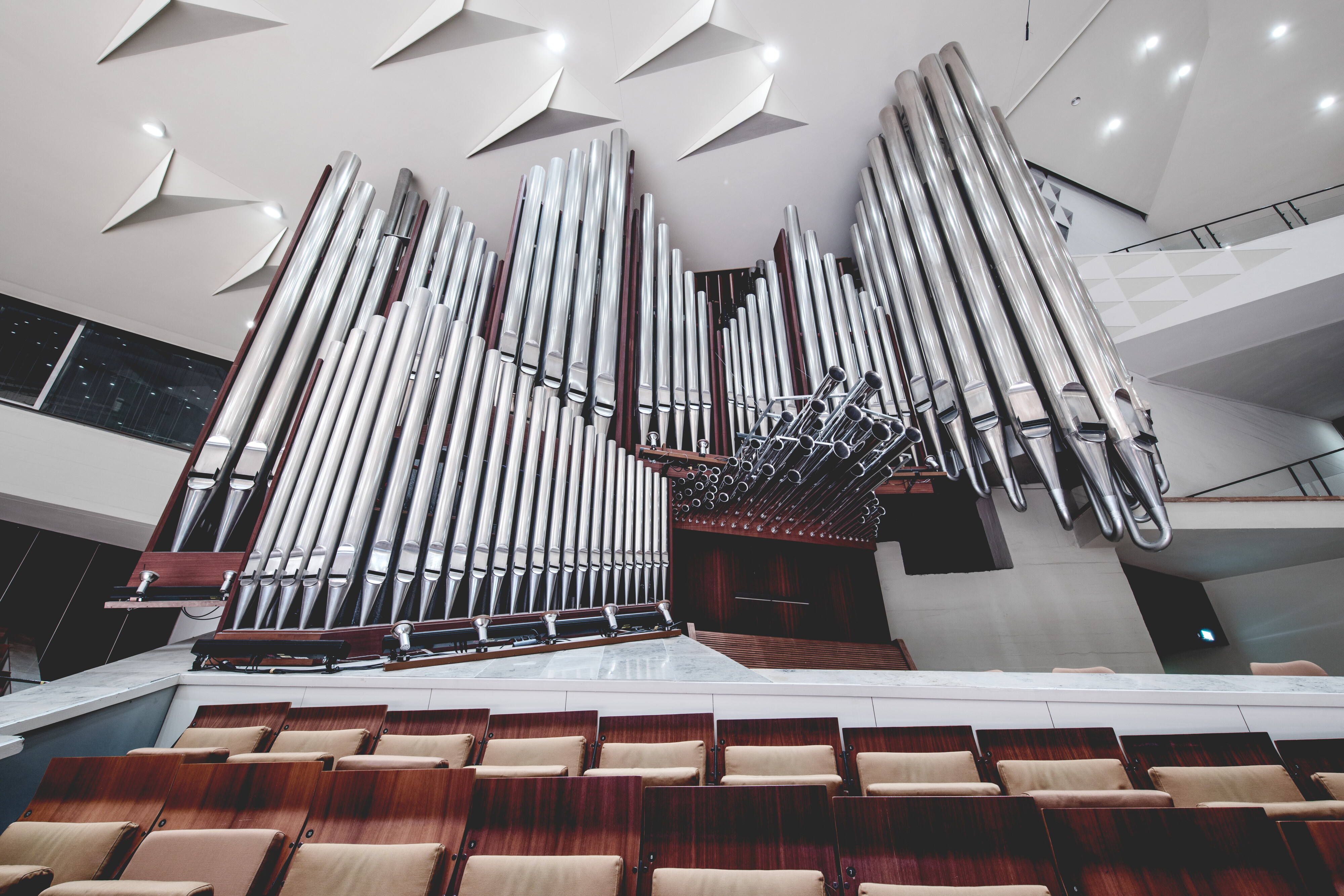 A large pipe organ with numerous shiny metal pipes is mounted at the front of a modern concert hall, with empty wooden seats and a white ceiling featuring geometric designs.