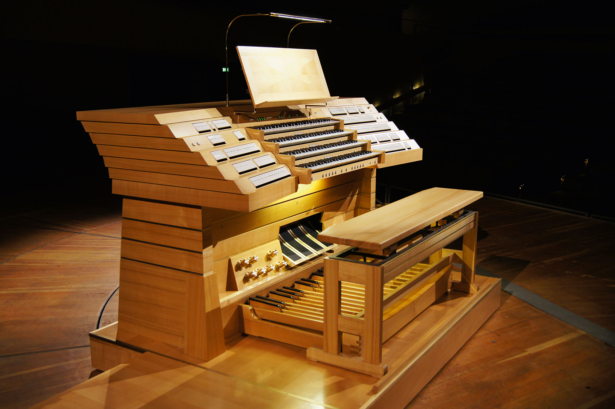 A modern wooden pipe organ console with multiple keyboards, numerous stop tabs, foot pedals, and a bench, illuminated by a lamp, set on a stage with a dark background.