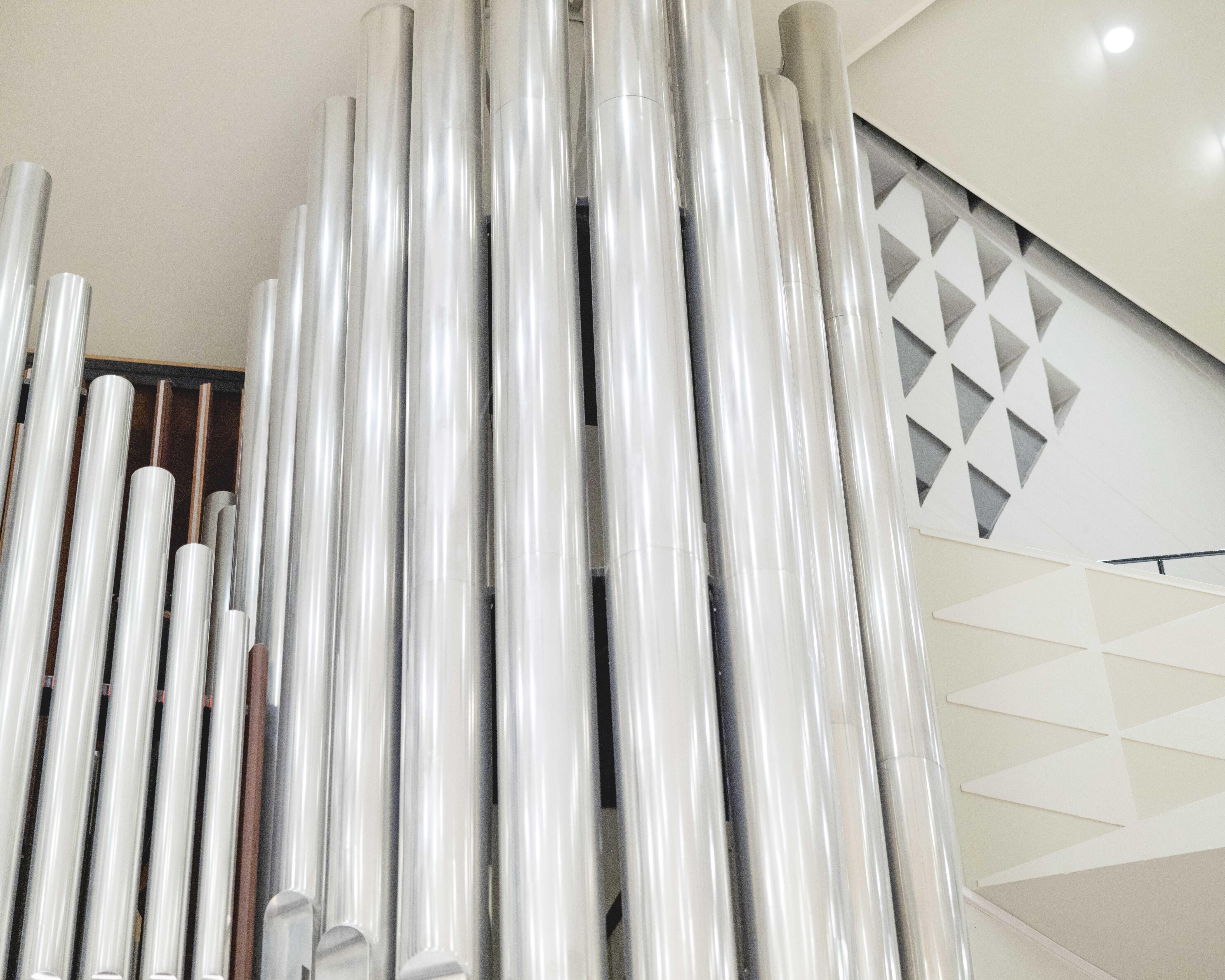 Große, silbern glänzende Orgelpfeifen stehen vertikal im Raum, mit geometrischen Wandmustern und heller Beleuchtung im Hintergrund, was eine moderne und klare architektonische Szene schafft.