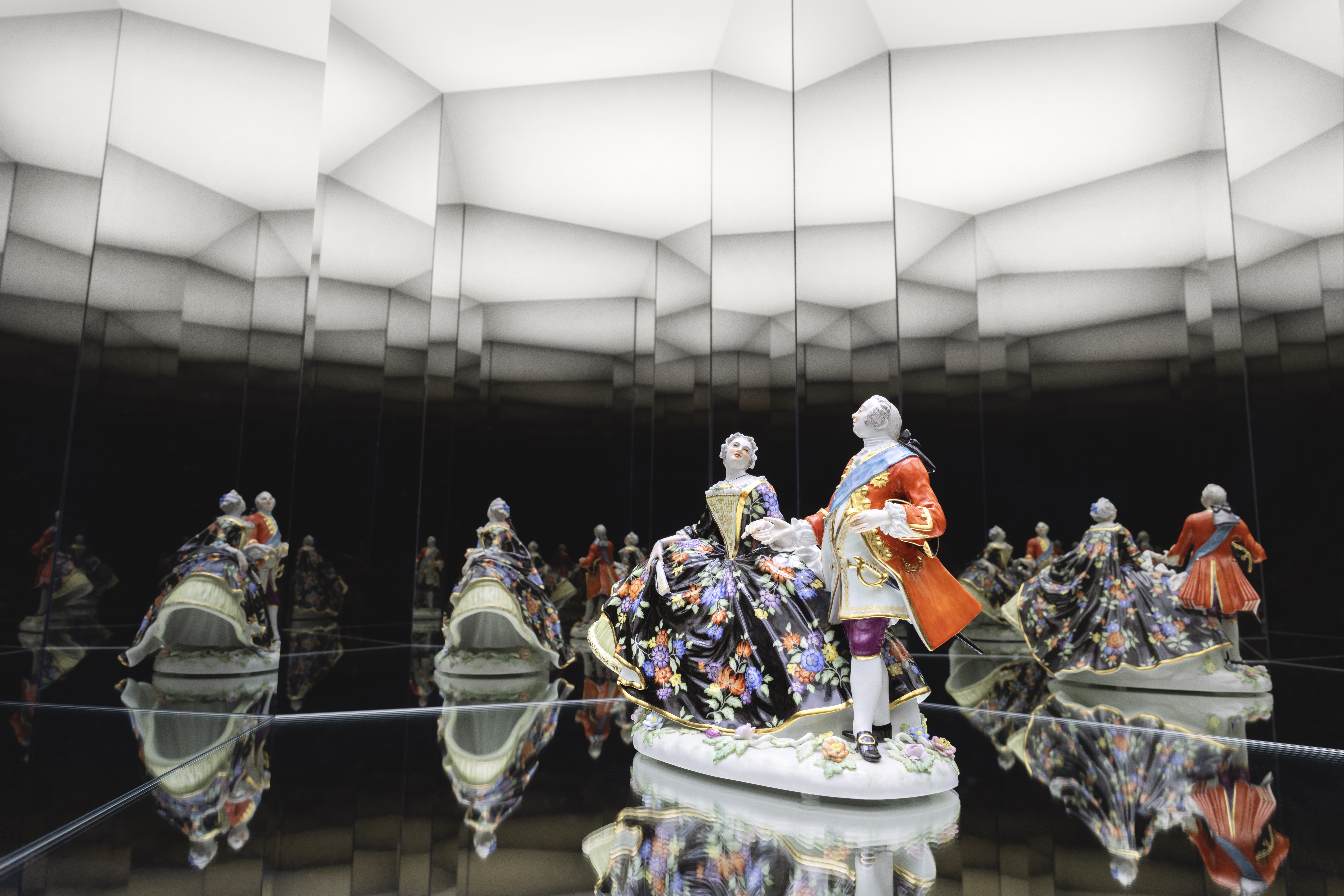 A porcelain figurine of a man and a woman in elaborate, historical clothing stands on a mirrored surface, which is reflected multiple times by the surrounding mirrors in a geometric, illuminated room.