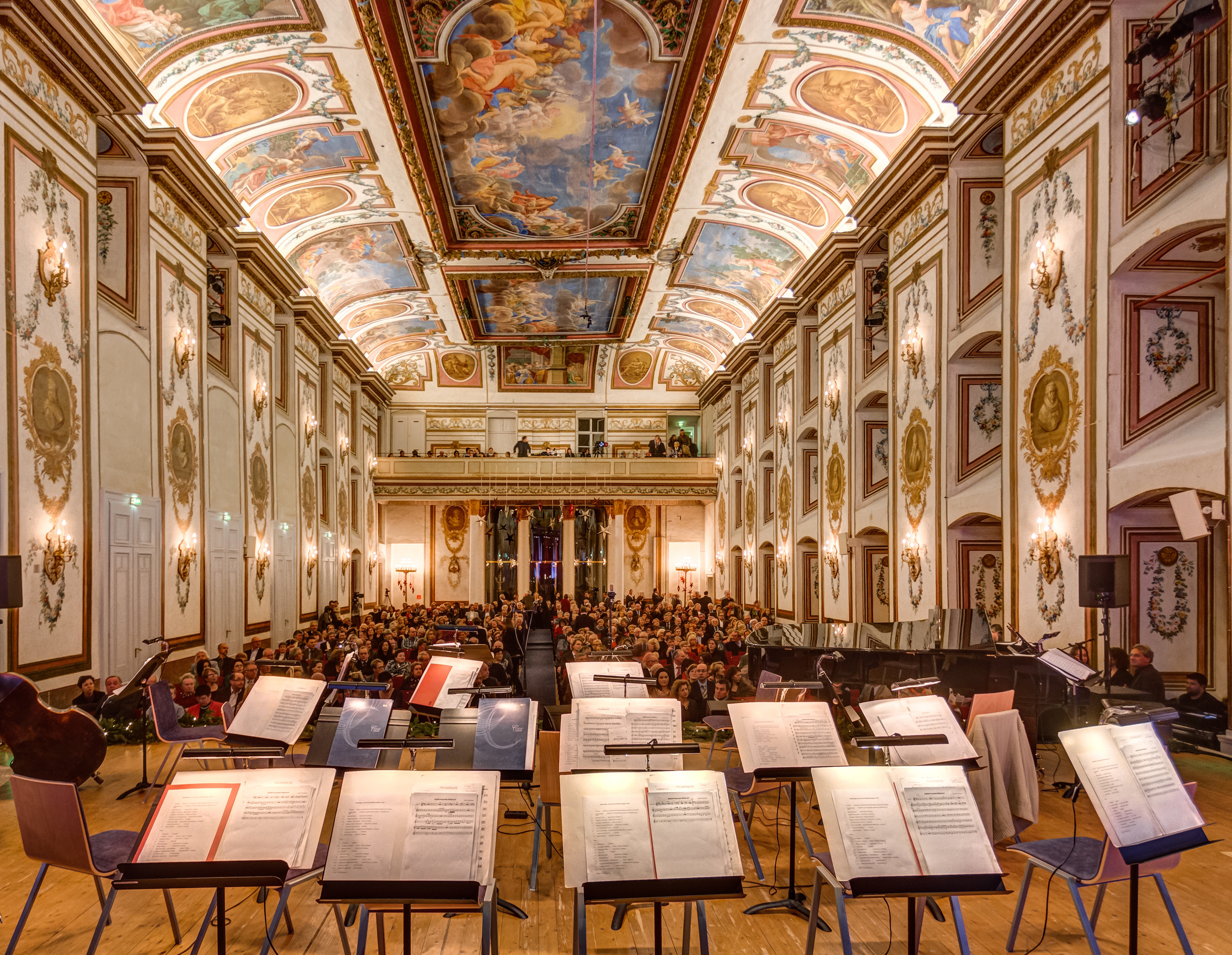 A grand, ornate concert hall with a painted ceiling, golden decorations, and an audience seated before empty music stands on stage, viewed from the performers’ perspective.
