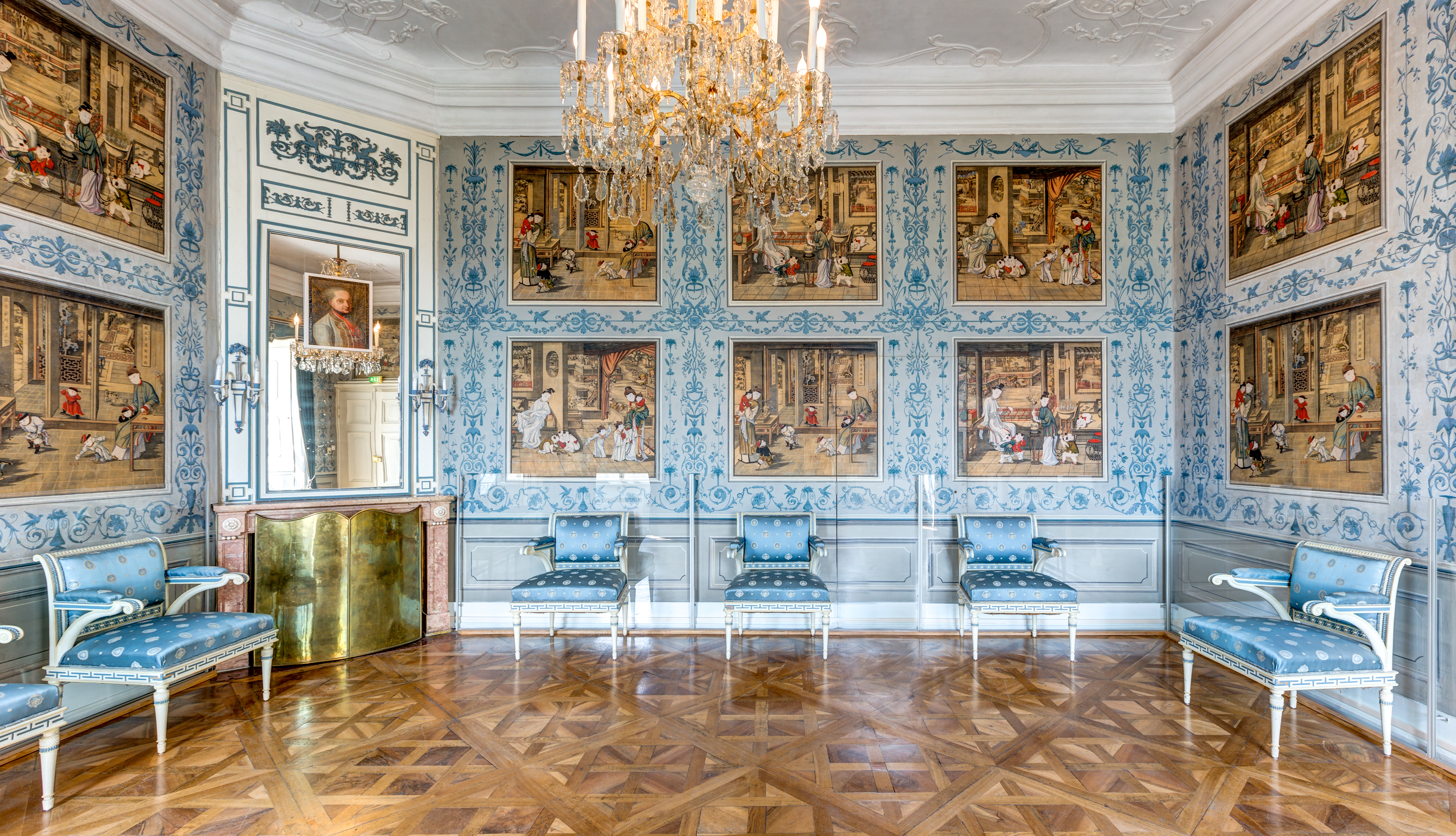 Elegant room with ornate blue-and-white walls featuring Chinese landscape paintings, blue upholstered chairs, a chandelier, and a patterned wood floor. A gold-framed mirror and portrait are above a gilded fireplace.