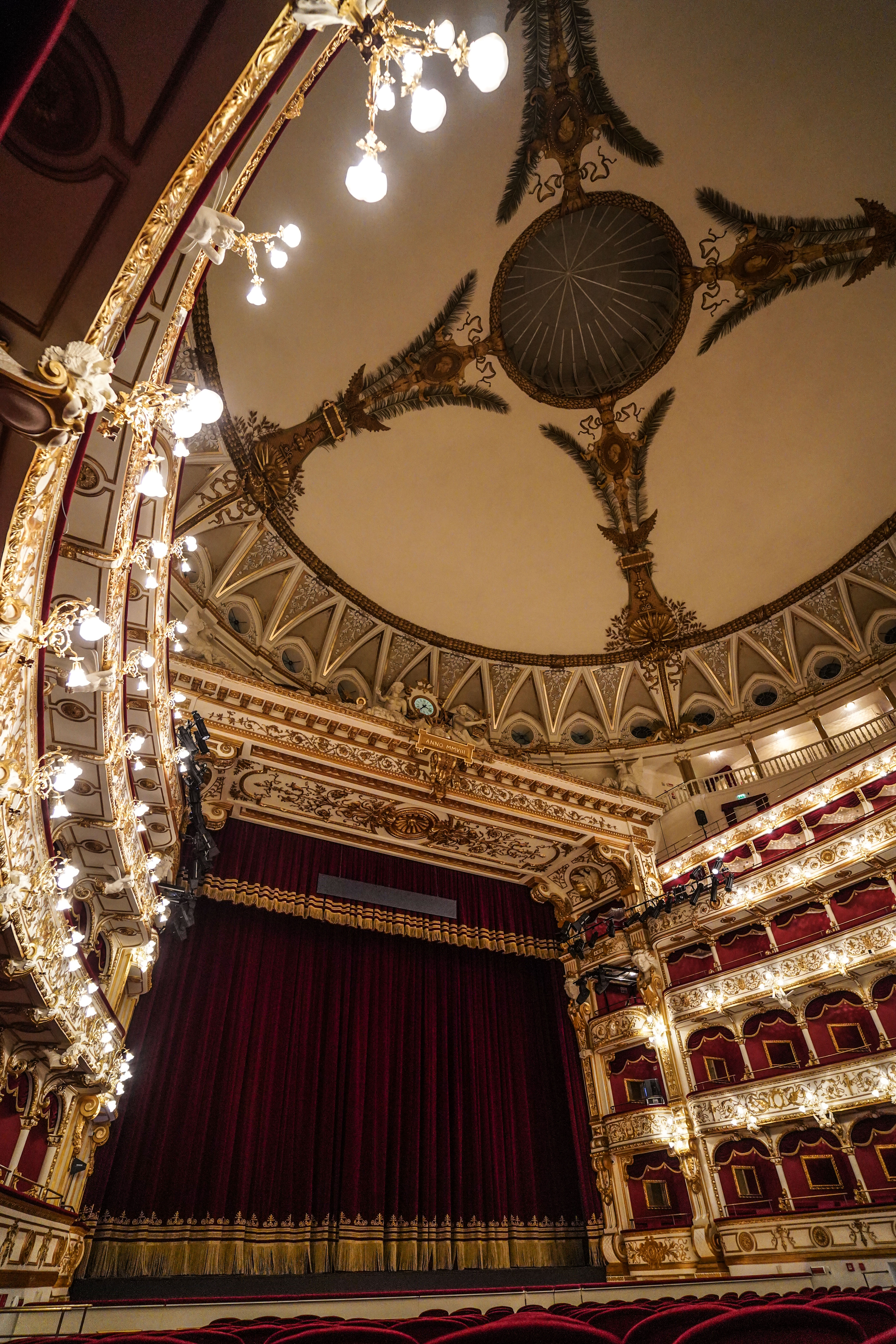 Elegant theatre interior with ornate gold decorations, red velvet seats, a large red stage curtain, several balconies and an ornate domed ceiling with decorative lighting and artistic details.
