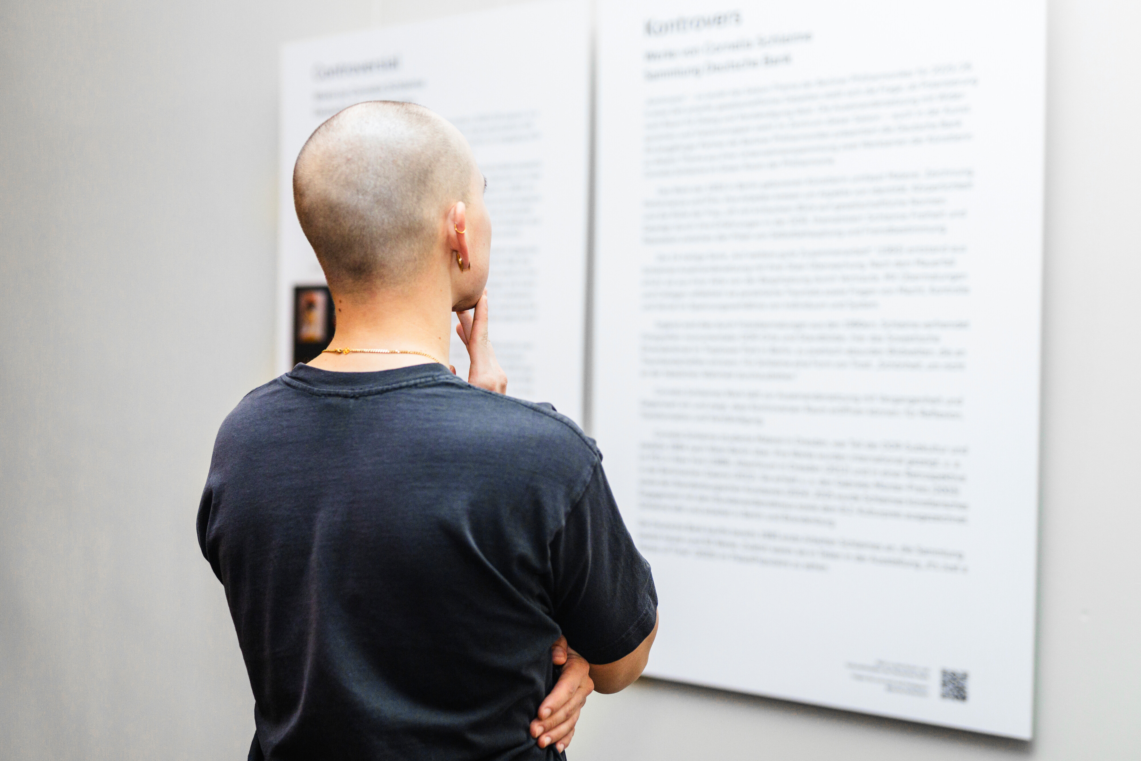 Eine Person mit rasiertem Kopf steht mit der Hand am Kinn da und liest nachdenklich die Texttafeln an einer Wand in einem Raum, der wie eine Galerie oder ein Museum aussieht.