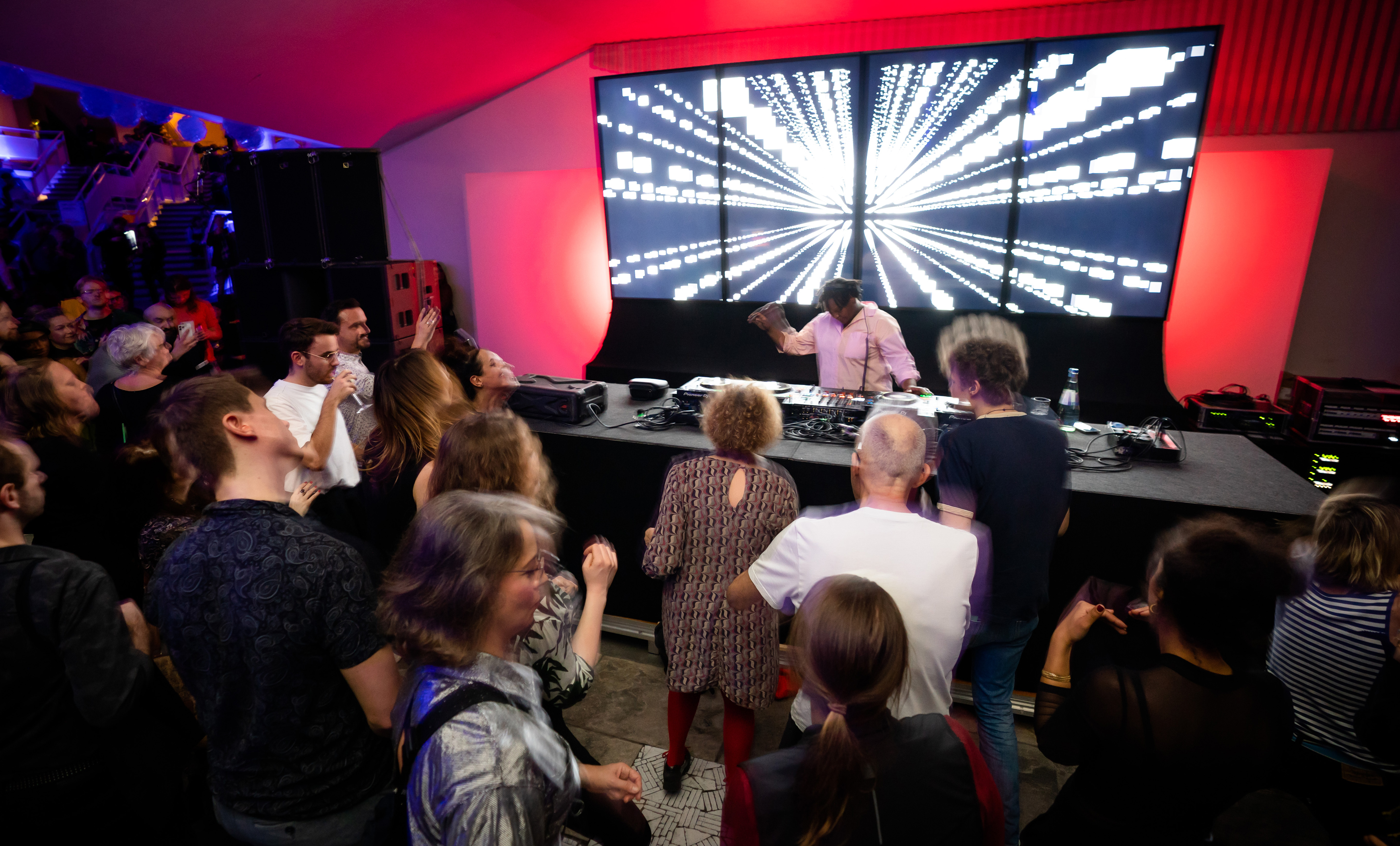 Eine DJ legt auf der Bühne auf, hinter ihm ein großes, leuchtendes Digitaldisplay. Davor versammelt sich eine Menschenmenge, tanzt und genießt die ausgelassene Atmosphäre. Der Raum ist rot und blau beleuchtet.