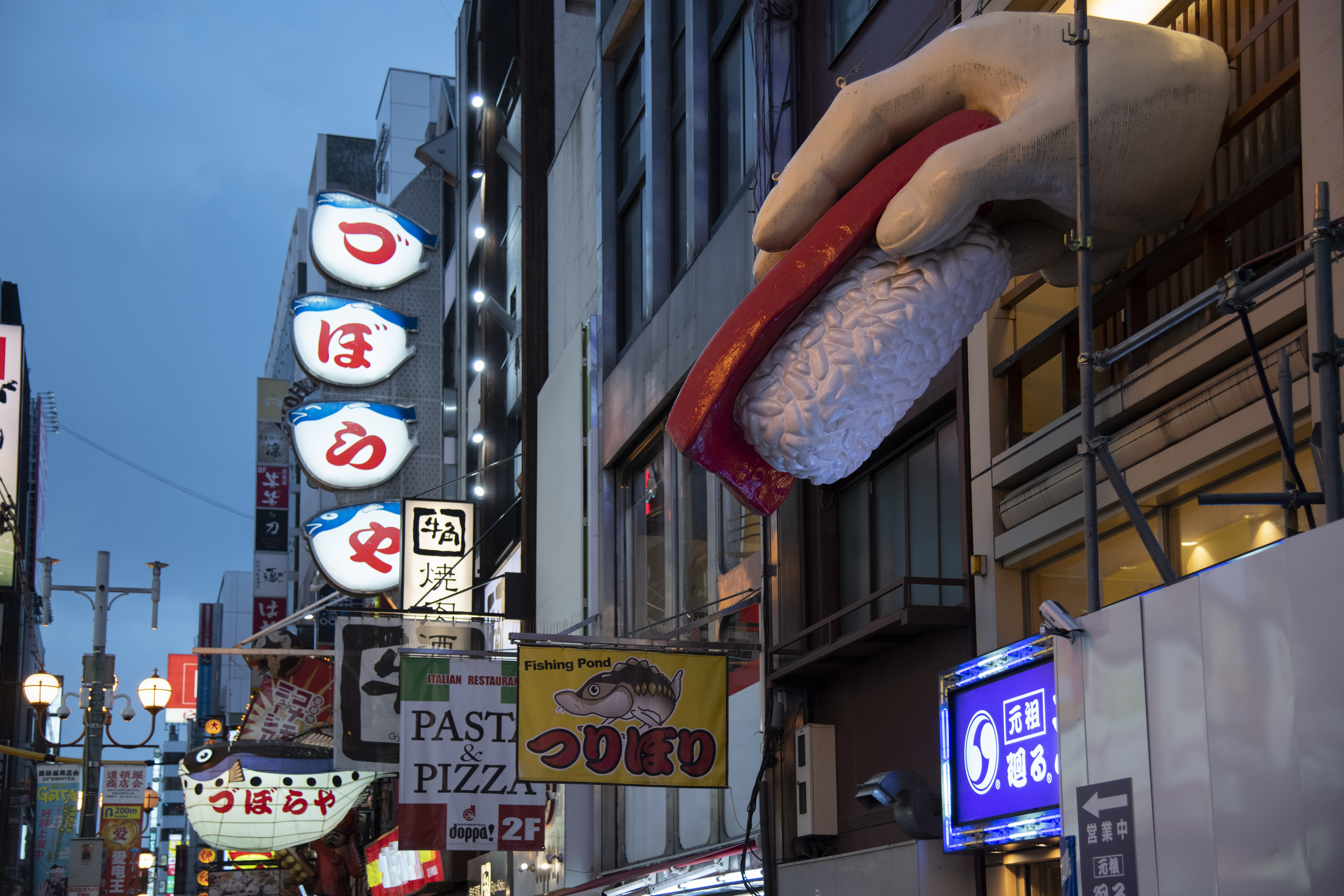 A city street in Japan at dusk shows colourful neon signs and a large sculpture of a hand holding sushi above a restaurant entrance. The buildings are densely packed, and various shop signs can be seen.