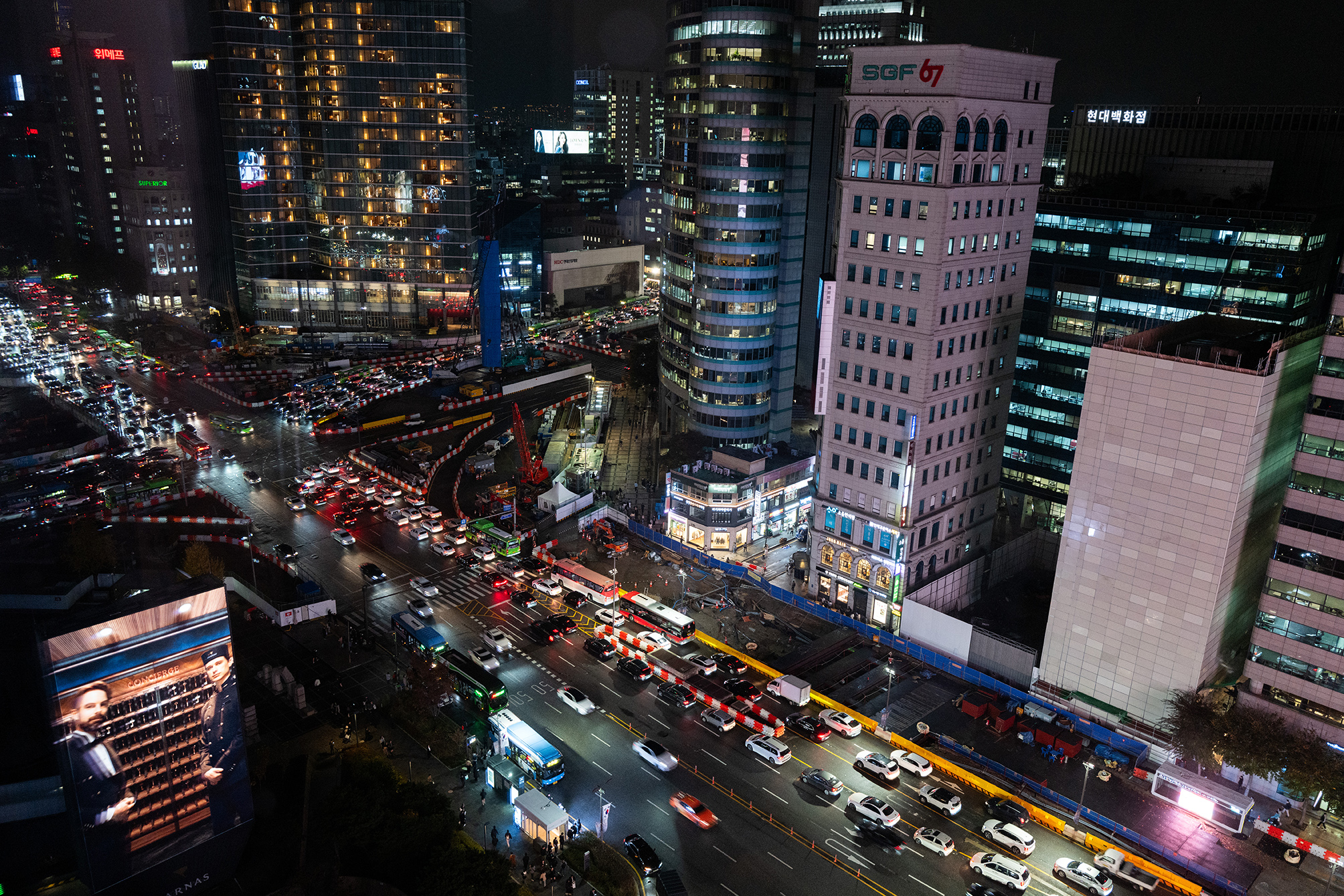 A busy city street at night filled with heavy traffic and illuminated buildings. Colorful lights and large digital billboards reflect off the wet roads, creating a vibrant urban scene.