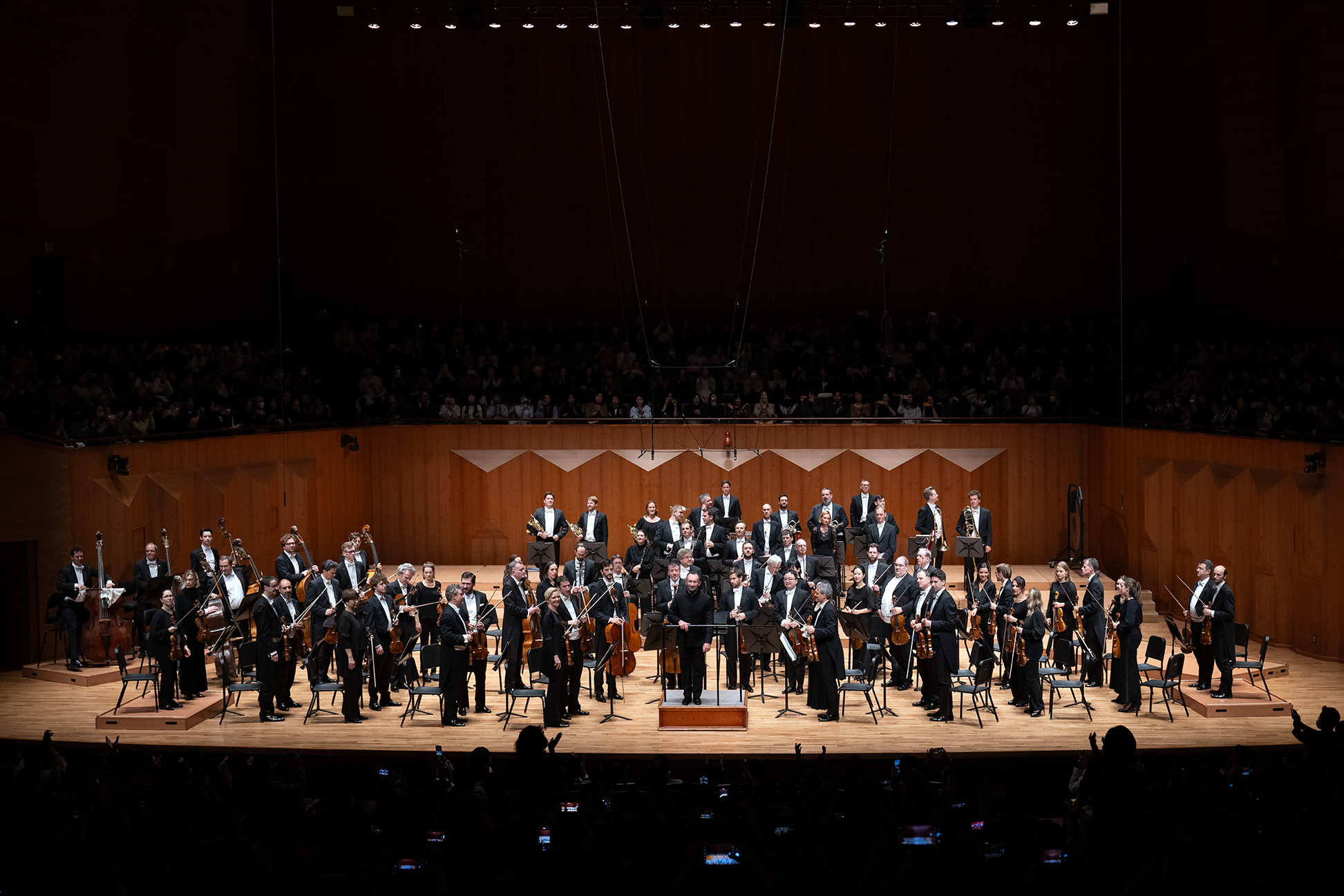 A full orchestra stands on stage, holding their instruments and facing the audience in a large concert hall with wooden walls. The audience is seated in the background.