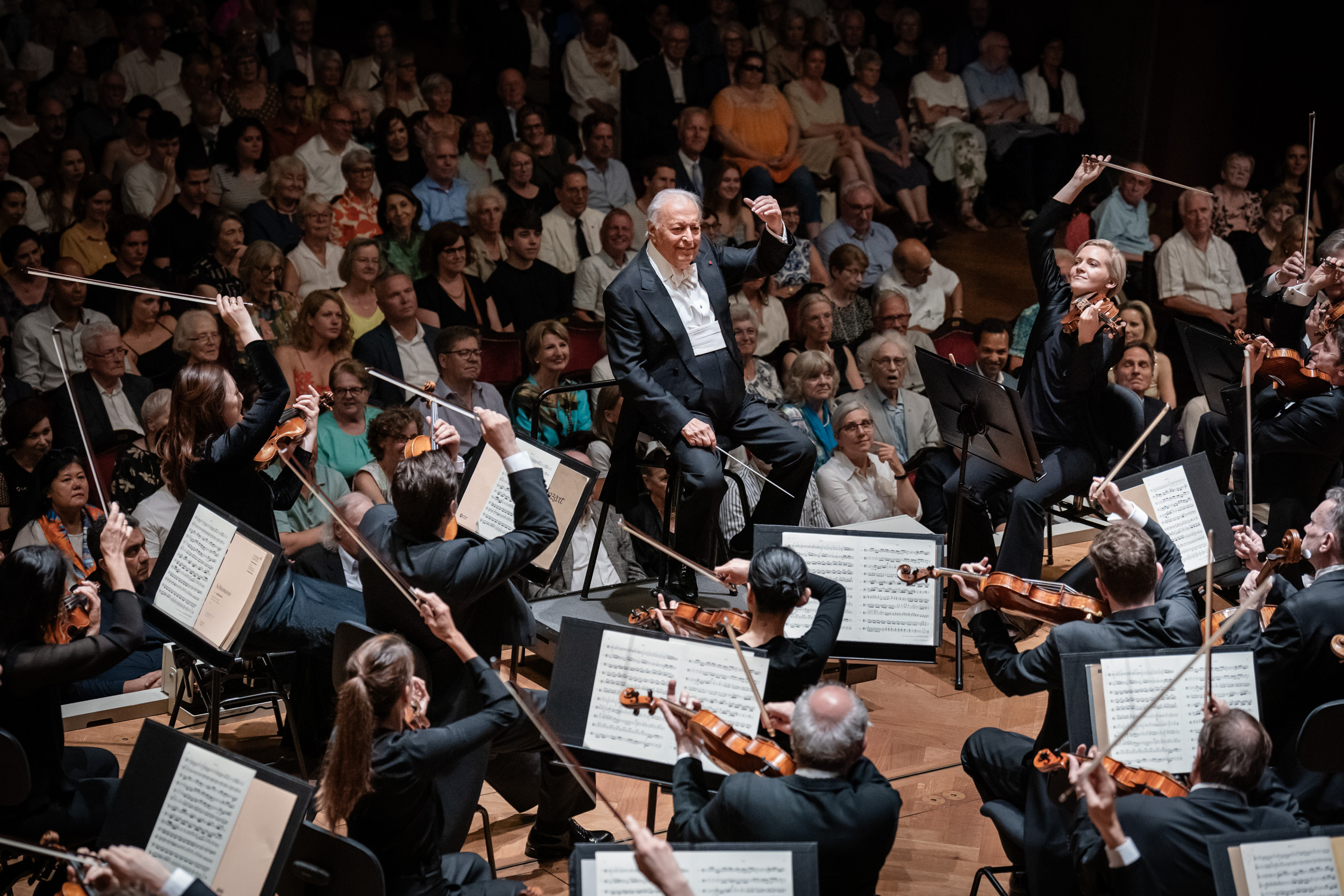 Ein Dirigent leitet ein Orchester auf der Bühne vor einem großen, aufmerksamen Publikum in einem Konzertsaal. Die Musiker halten Geigen und Celli und lesen von den Notenblättern ab.