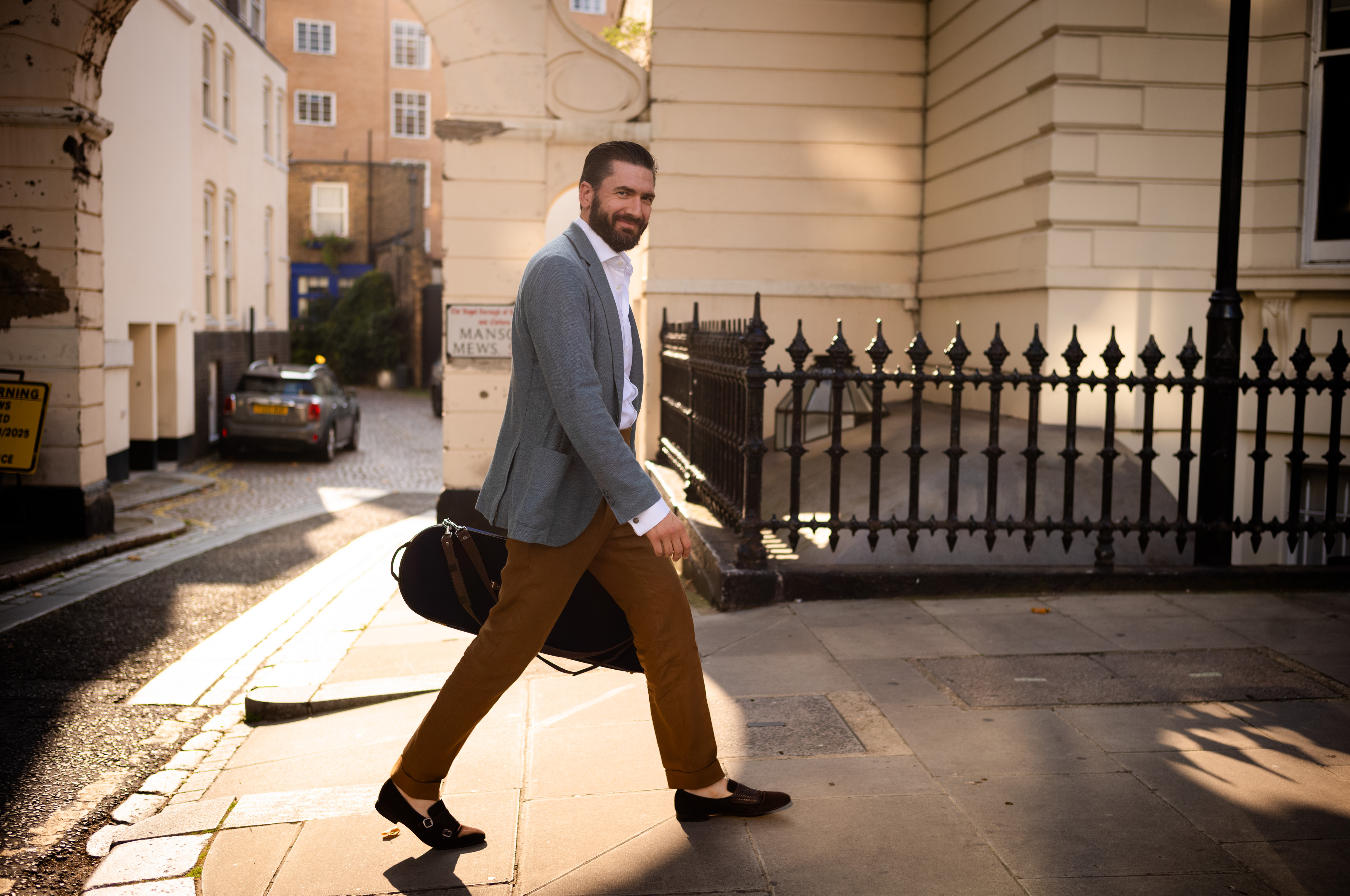 Dorian Xhoxhi with a beard, a grey blazer and brown trousers is walking on a sunlit pavement in the city, carrying a black guitar case. Buildings and a parked car can be seen in the background.