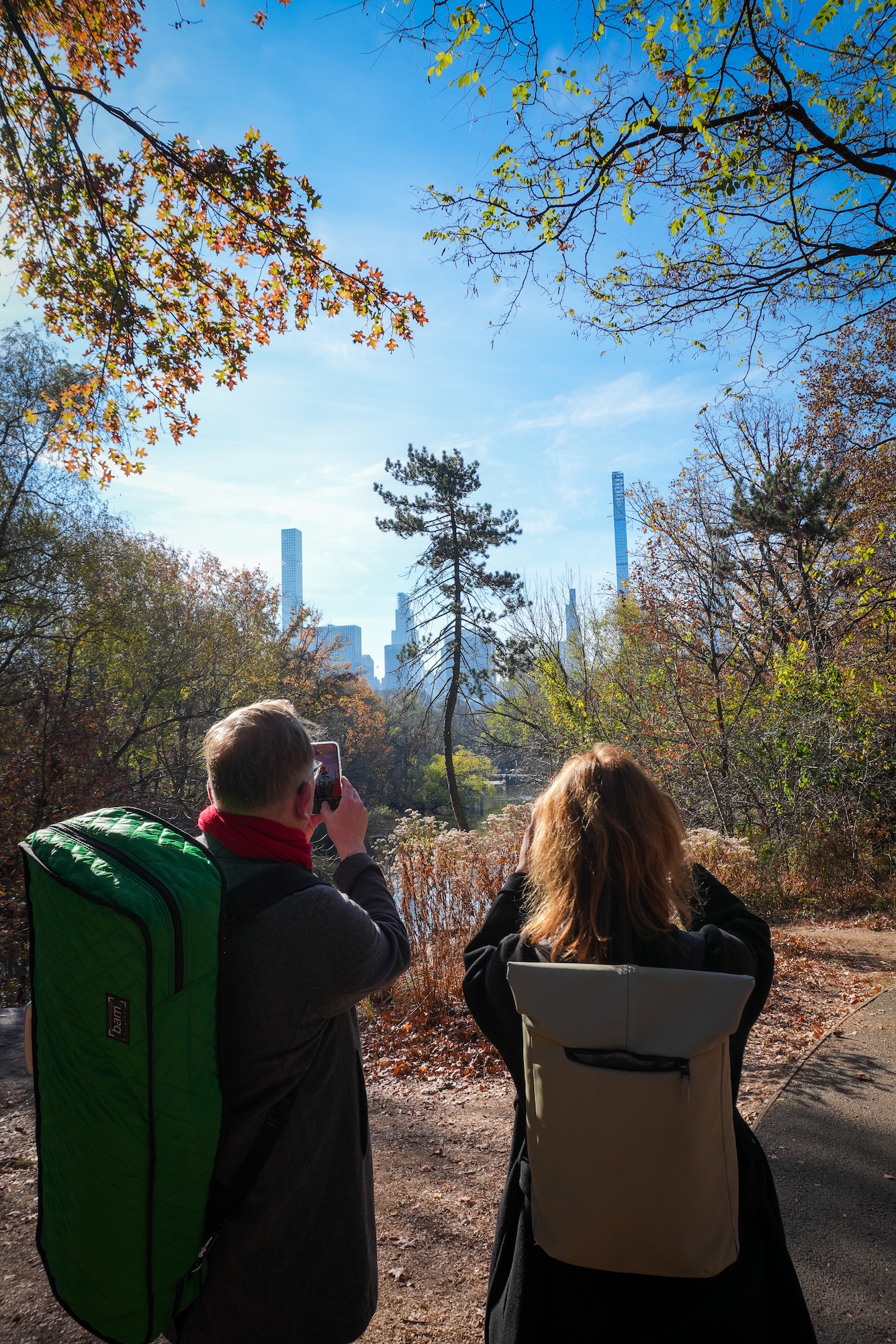 Two people with rucksacks photograph the skyline of a city from a wooded park path on a sunny day, surrounded by autumn trees with colourful leaves under a clear blue sky.