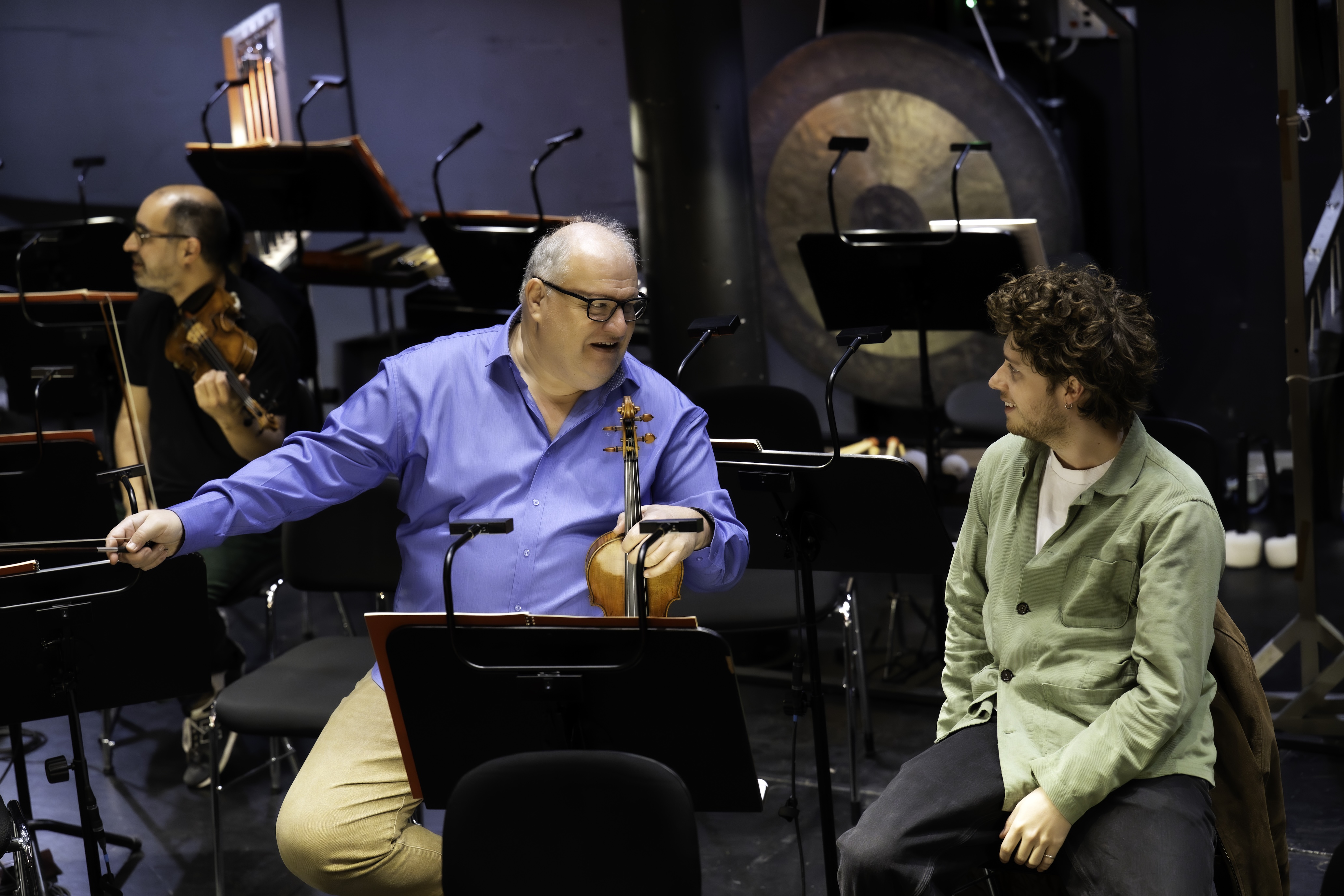 Zwei Geiger sitzen in einem Orchester und unterhalten sich. Im Hintergrund sind Notenständer und Instrumente zu sehen.