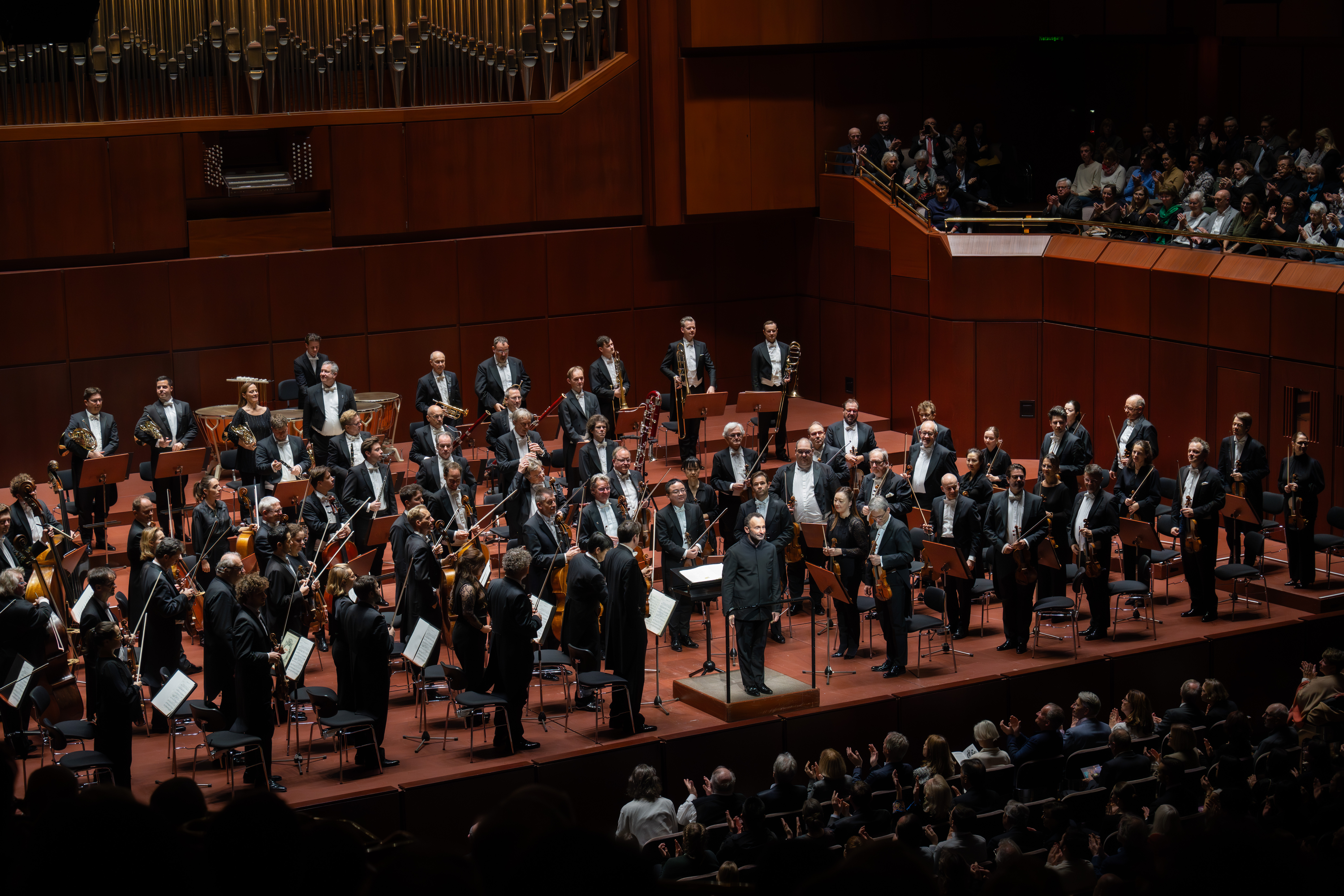 Die Berliner Philharmoniker stehen mit ihrem festlich gekleideten Dirigenten auf der Bühne eines großen Konzertsaals, während das Publikum zuschaut und applaudiert.
