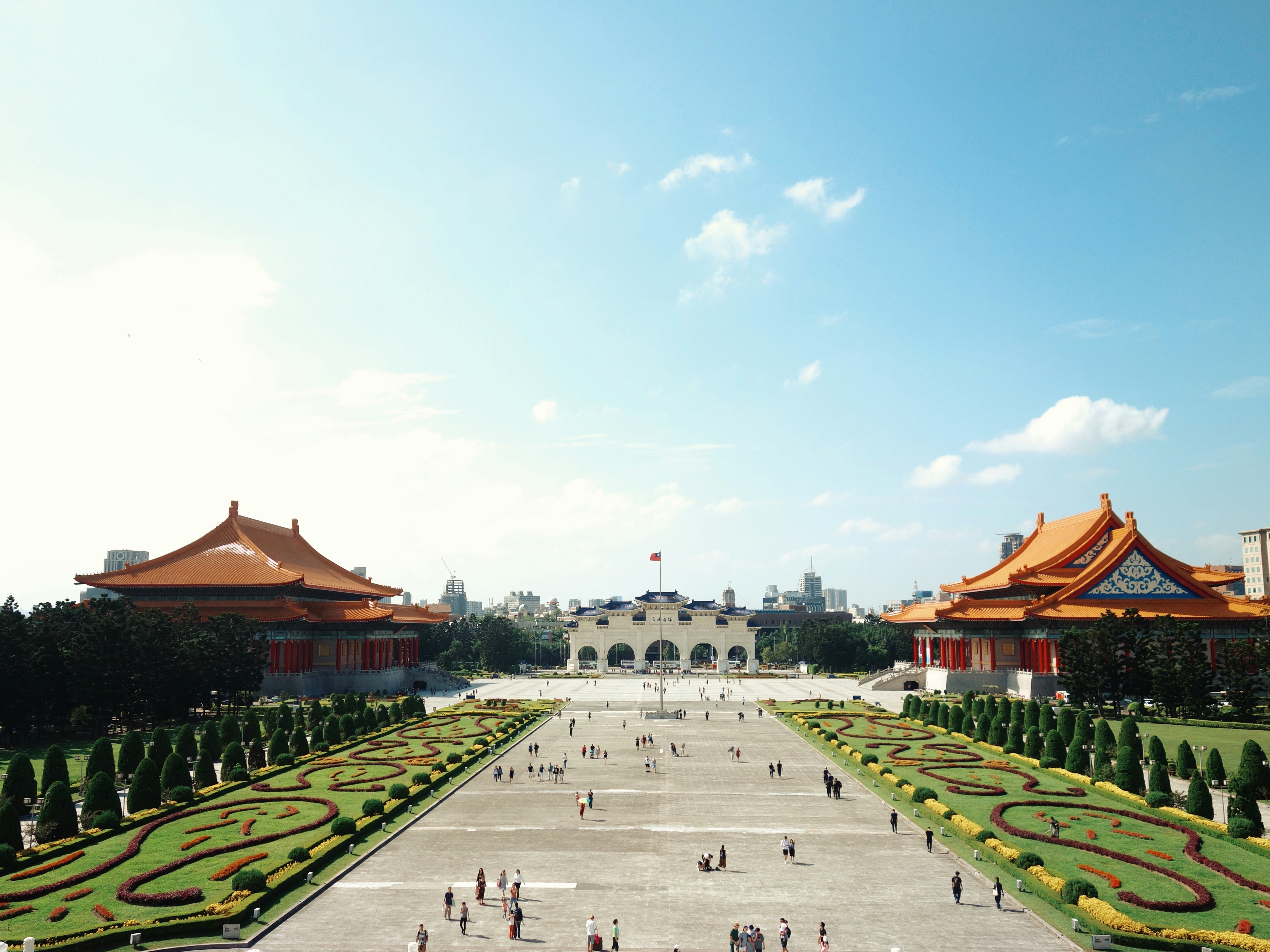 Ein breiter Platz mit dekorativen Gärten und beschnittenen Bäumen führt zu einem weißen Torbogen; zwei große traditionelle chinesische Gebäude mit orangefarbenen Dächern stehen auf beiden Seiten unter einem blauen Himmel mit vereinzelten Wolken.