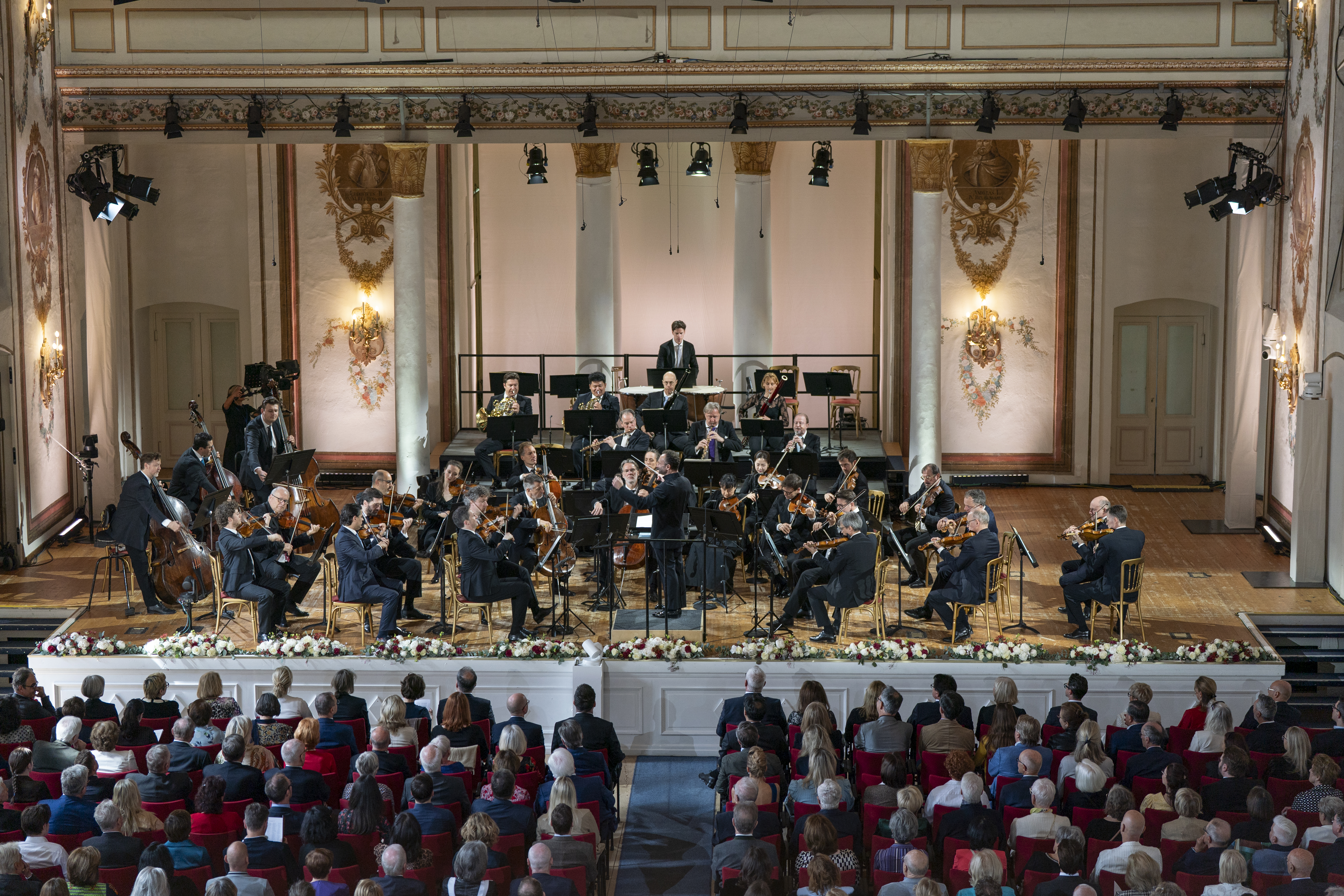 Ein Orchester spielt auf einer geschmückten Bühne in einem großen Konzertsaal. Die Musiker sitzen in einem Halbkreis und der Dirigent steht in der Mitte. Das Publikum sitzt in Reihen roter Stühle und schaut zu.