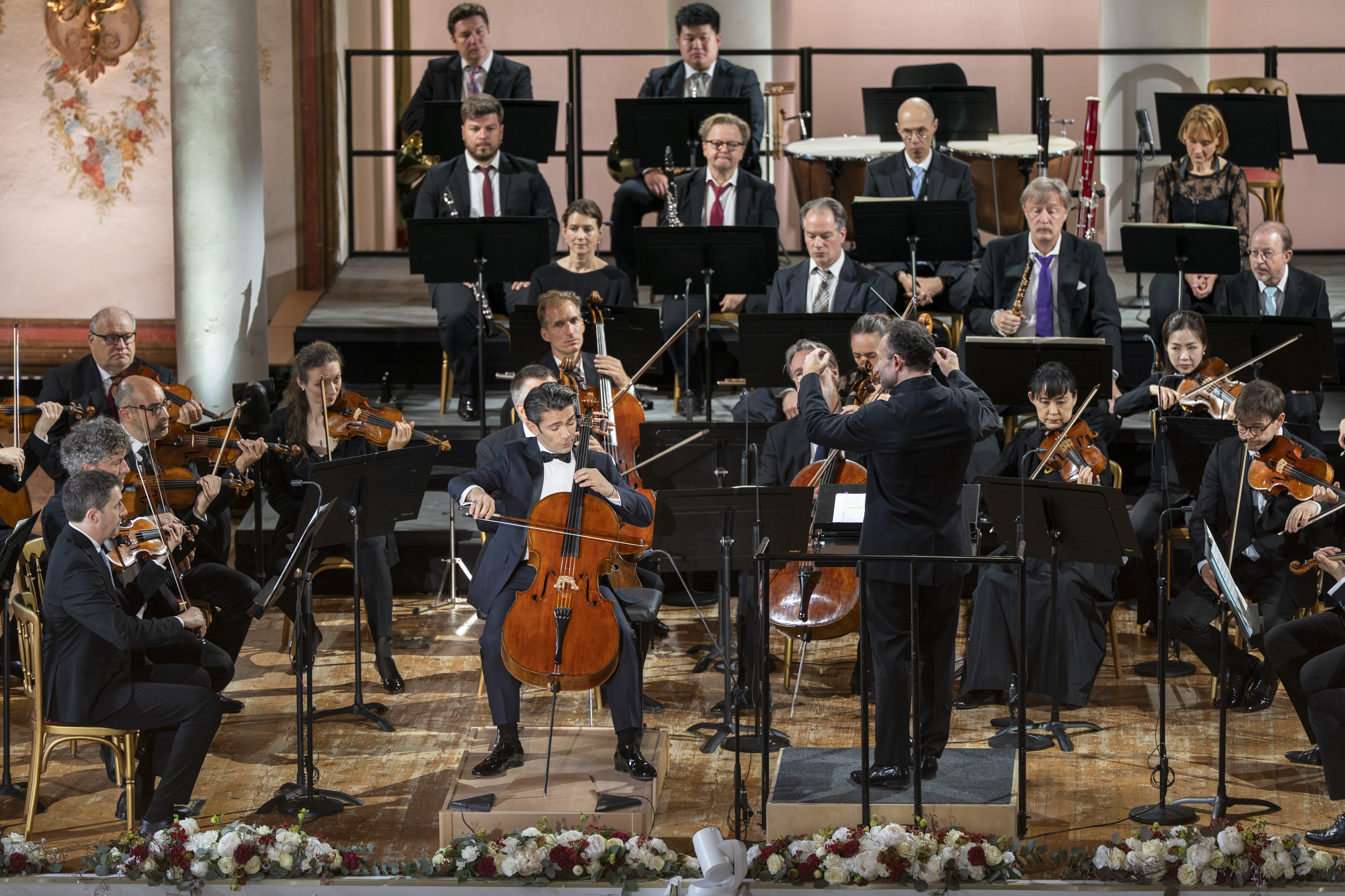 Nahaufnahme der Bühne, auf der der Solist Gautier Capuçon mit seinem Violoncello spielt, Chefdirigent Kirill Petrenko dirigiert, die beiden sind umgeben vom spielenden Orchester.
