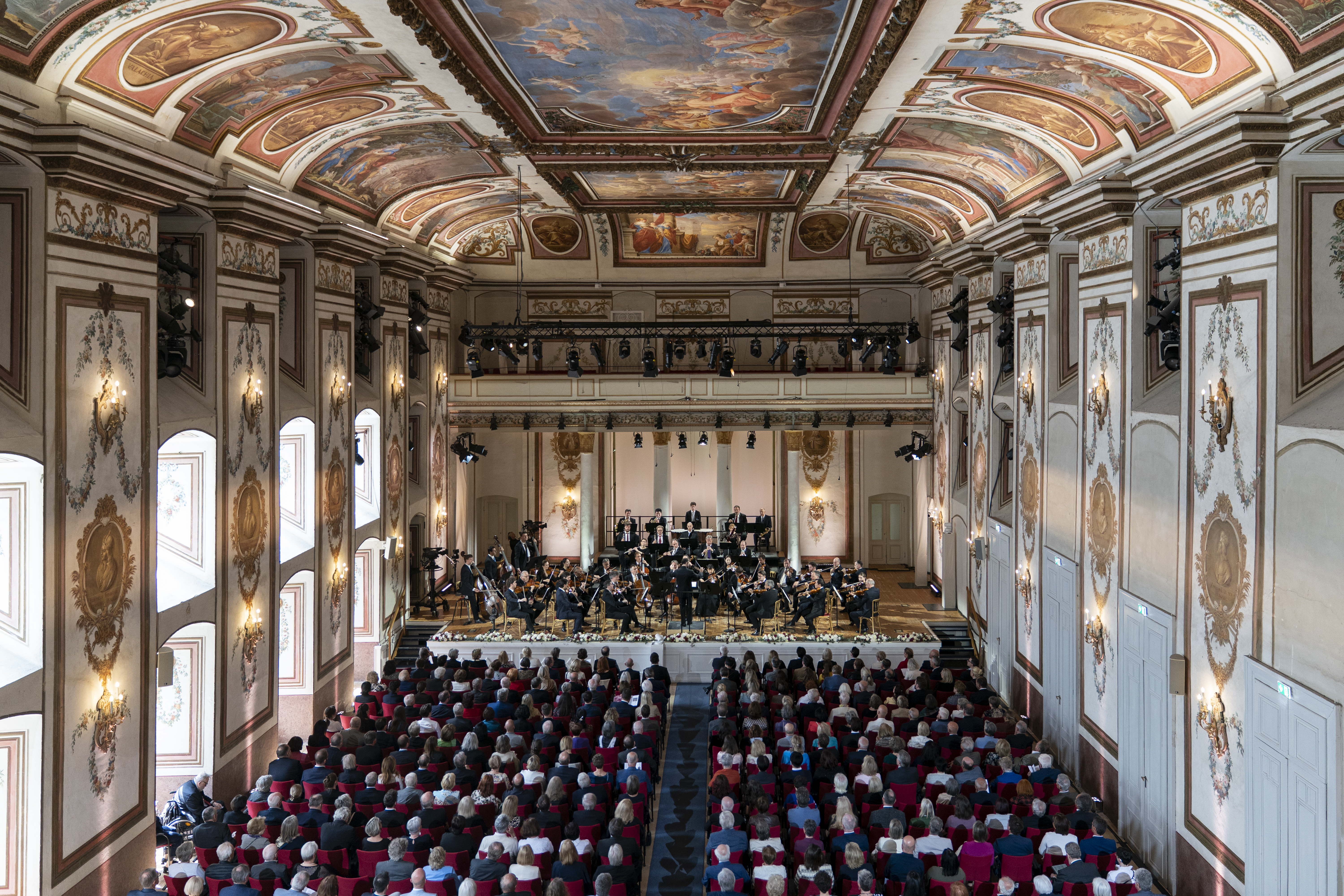 Ein großes Publikum verfolgt die Darbietung eines Symphonieorchesters auf der Bühne eines prunkvollen Konzertsaals mit hohen Decken und kunstvollen Wand- und Deckendekorationen.