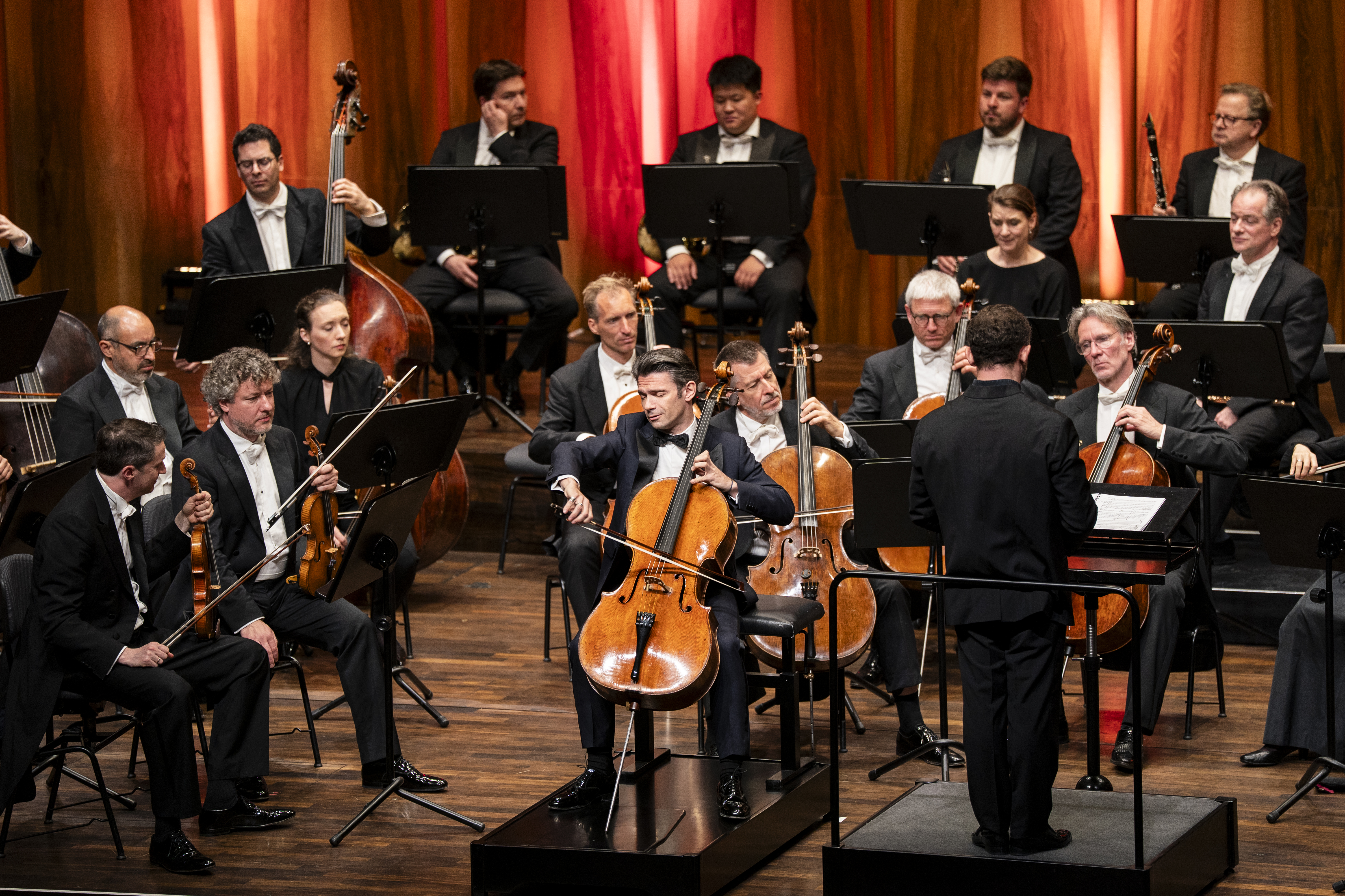 Nahaufnahme eines Orchesters in Aktion. Cellist Gautier Capuçon steht im Zentrum und spielt konzentriert ein Solo, während Dirigent Kirill Petrenko rechts im Bild steht. Die Musiker*innen tragen klassische schwarze Abendgarderobe vor dem Hintergrund der warm beleuchteten Holzwand.