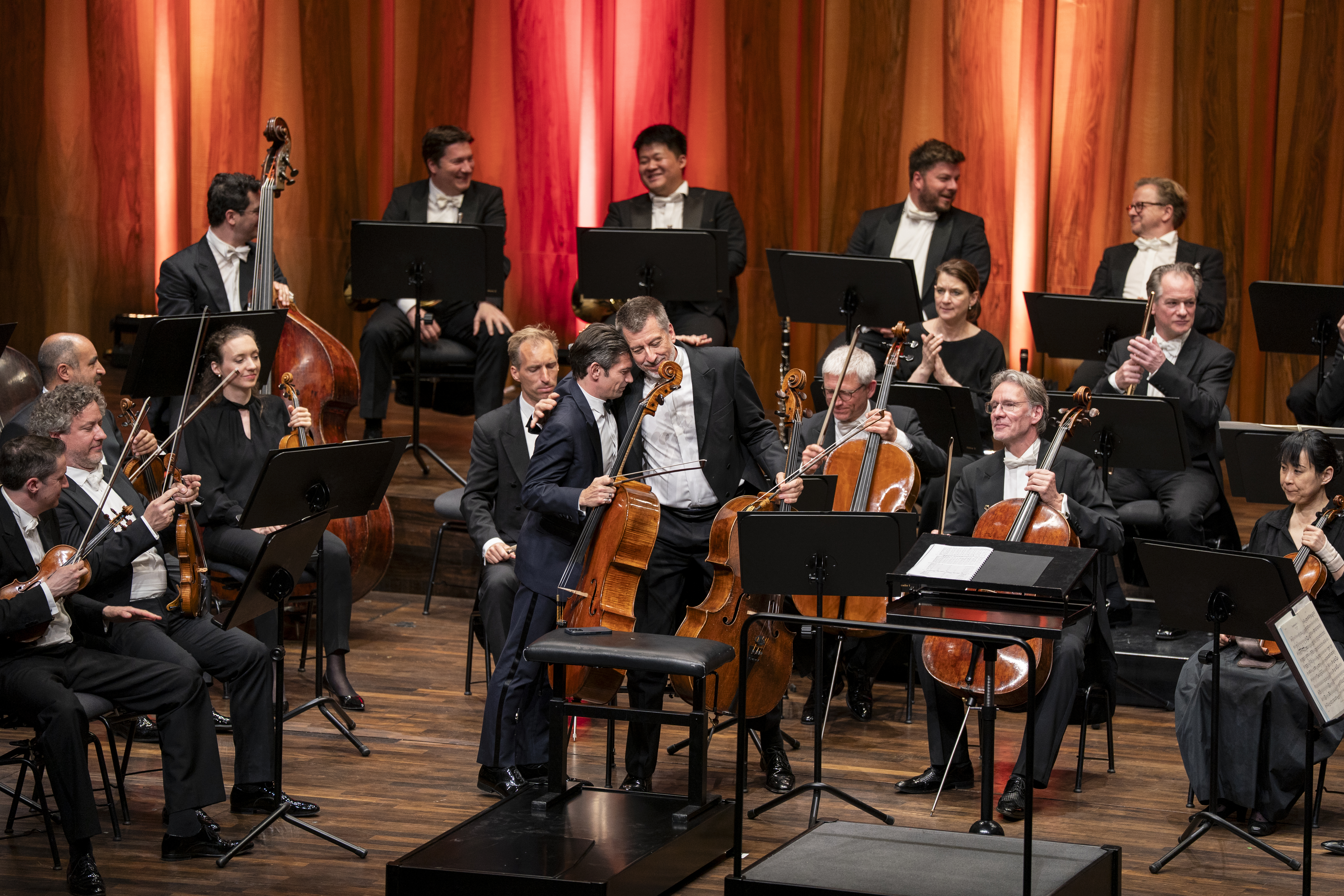 Cellisten Gautier Capuçon und Ludwig Quandt stehen auf der Bühne und umarmen sich herzlich. Um sie herum sind weitere Orchestermitglieder zu sehen, die Instrumente halten. Die Atmosphäre wirkt kollegial und gelöst.