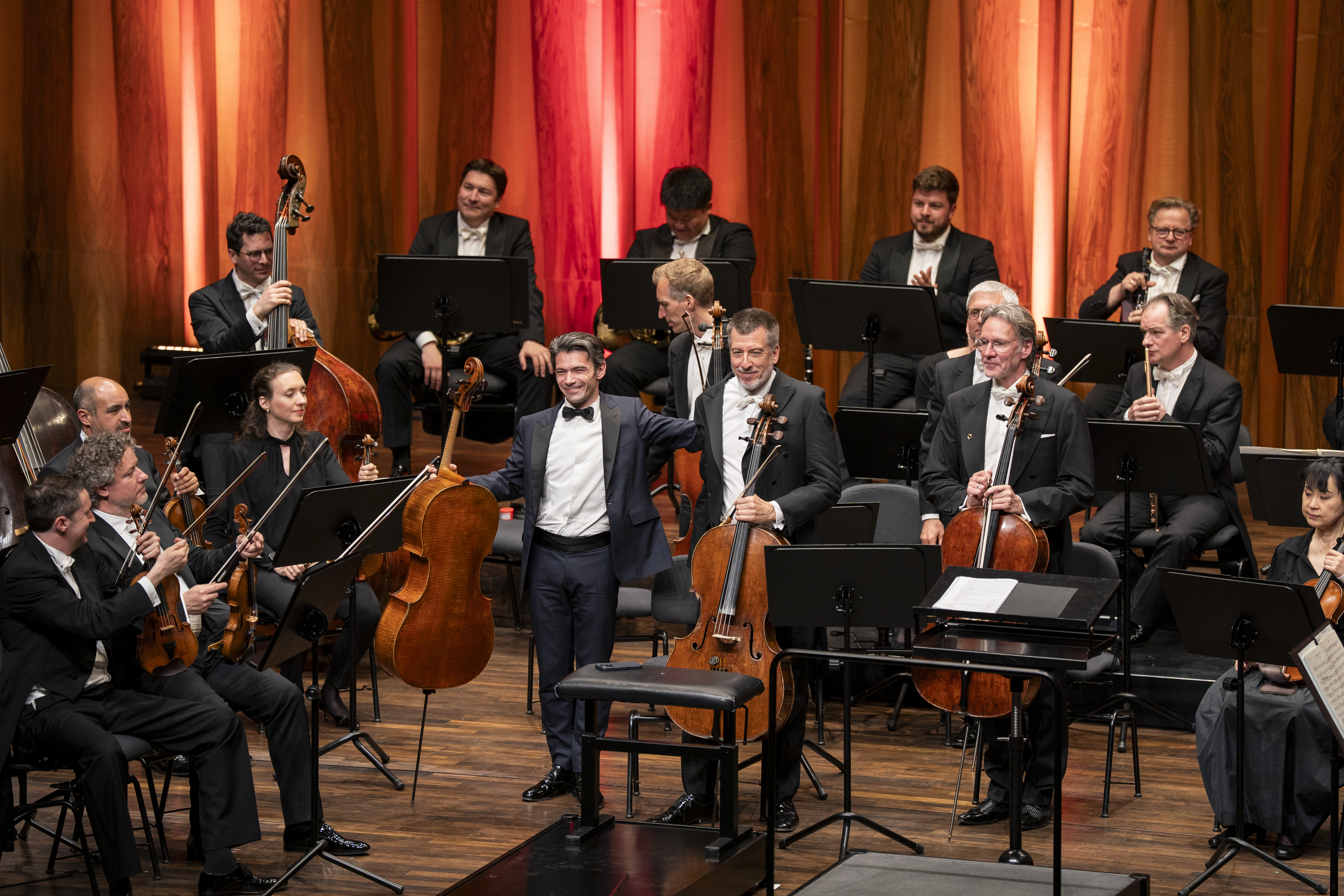 Cellist Gautier Capuçon steht lächelnd neben Solocellist Ludwig Quandt auf der Bühne und legt ihm freundschaftlich den Arm um die Schulter. Beide halten ihre Instrumente. Im Hintergrund sind die Bläser und Kontrabassisten des Orchesters zu sehen, die den Moment beobachten.