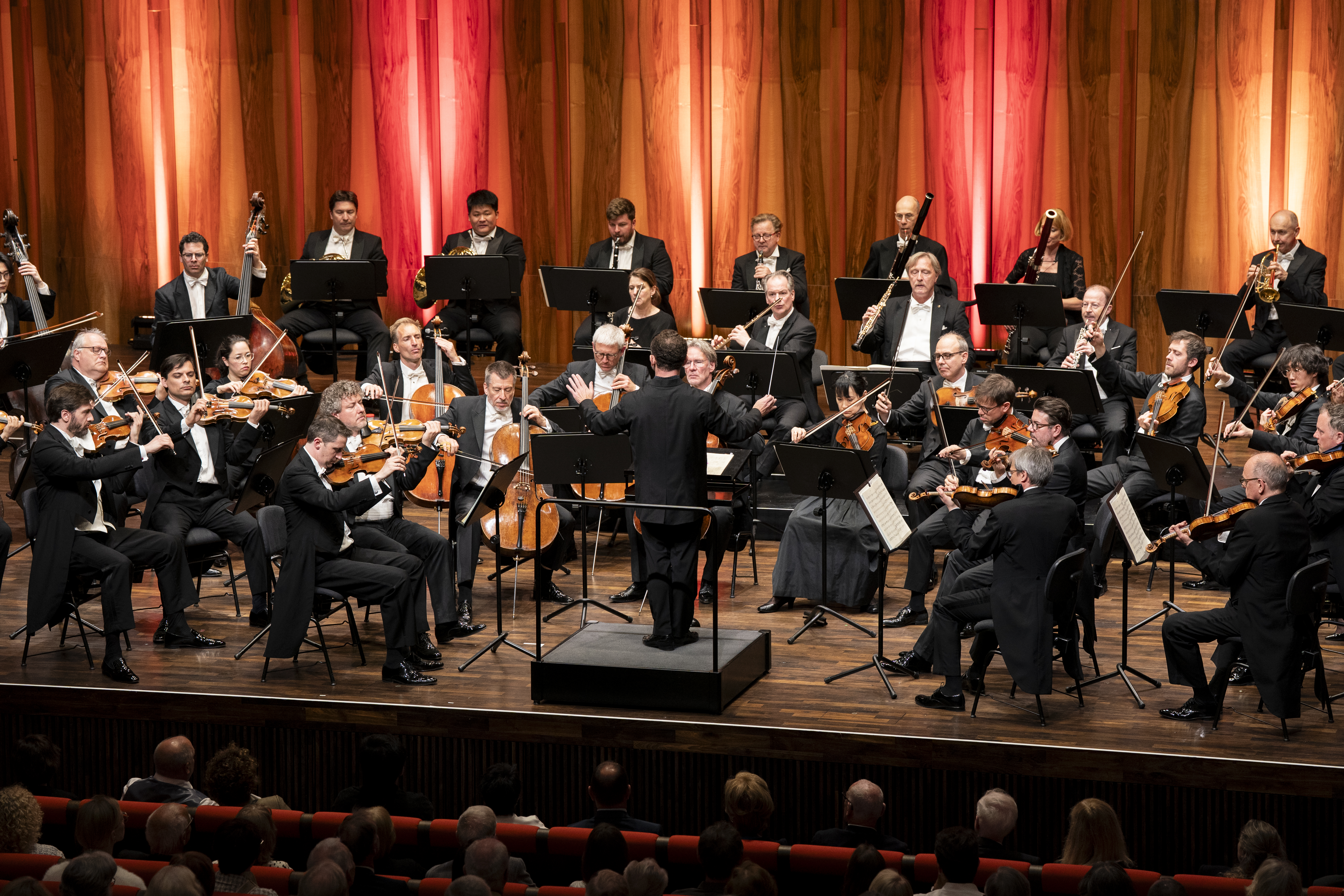 Das gesamte Orchester auf der Bühne unter der Leitung des Dirigenten Kirill Petrenko. Die Streicher im Vordergrund sind im vollen Spiel begriffen. Die vertikale Holzmaserung der Saalwand und die rote Beleuchtung erzeugen eine warme, fokussierte Akustik-Optik.