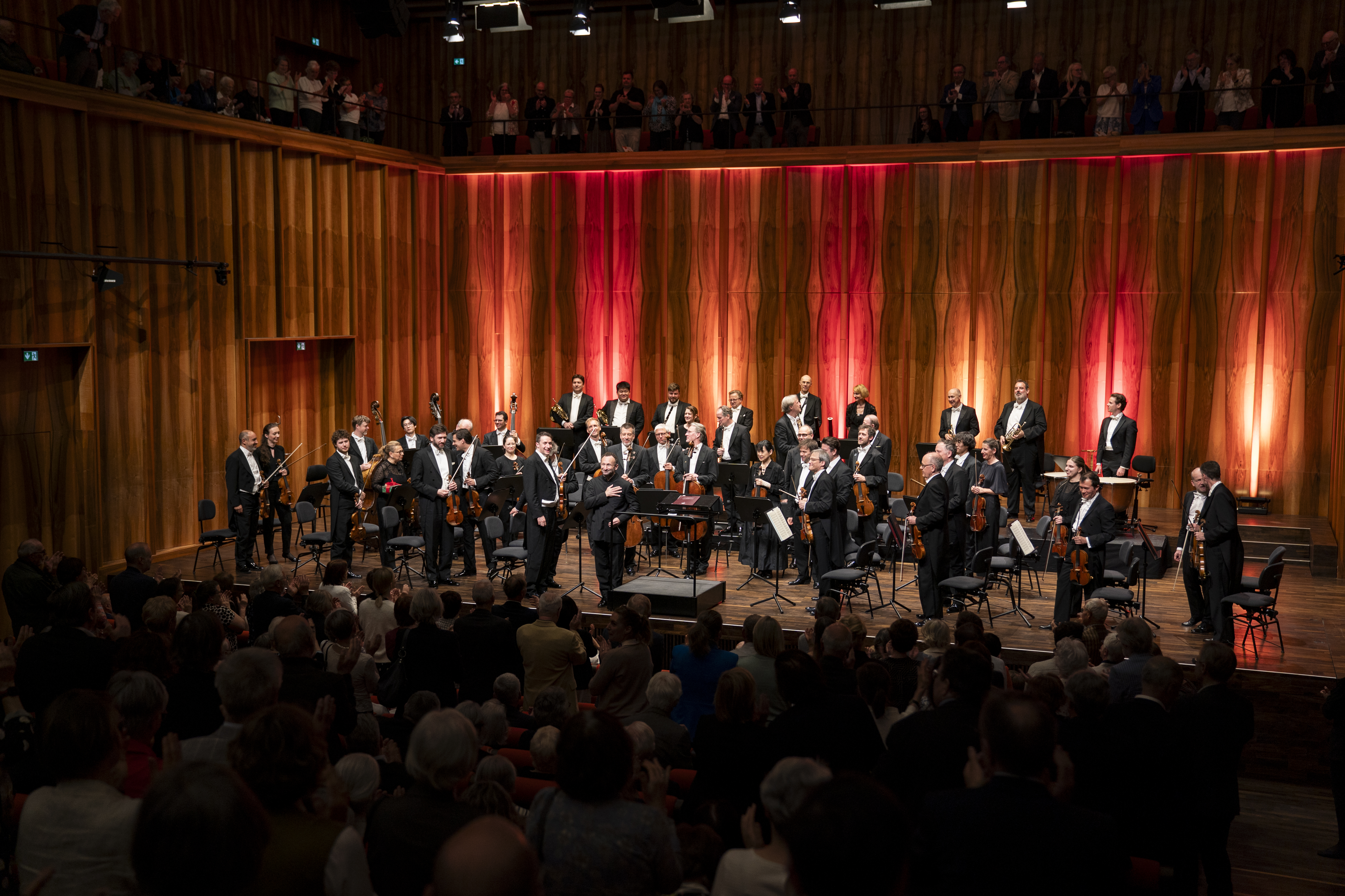 Das Orchester und der Dirigent stehen auf der Bühne und nehmen den Applaus des Publikums entgegen. Der Saal ist bis auf den letzten Platz besetzt, auch auf der Empore im Hintergrund stehen Menschen und klatschen. Die rote Beleuchtung der Holzwand setzt einen starken visuellen Akzent.