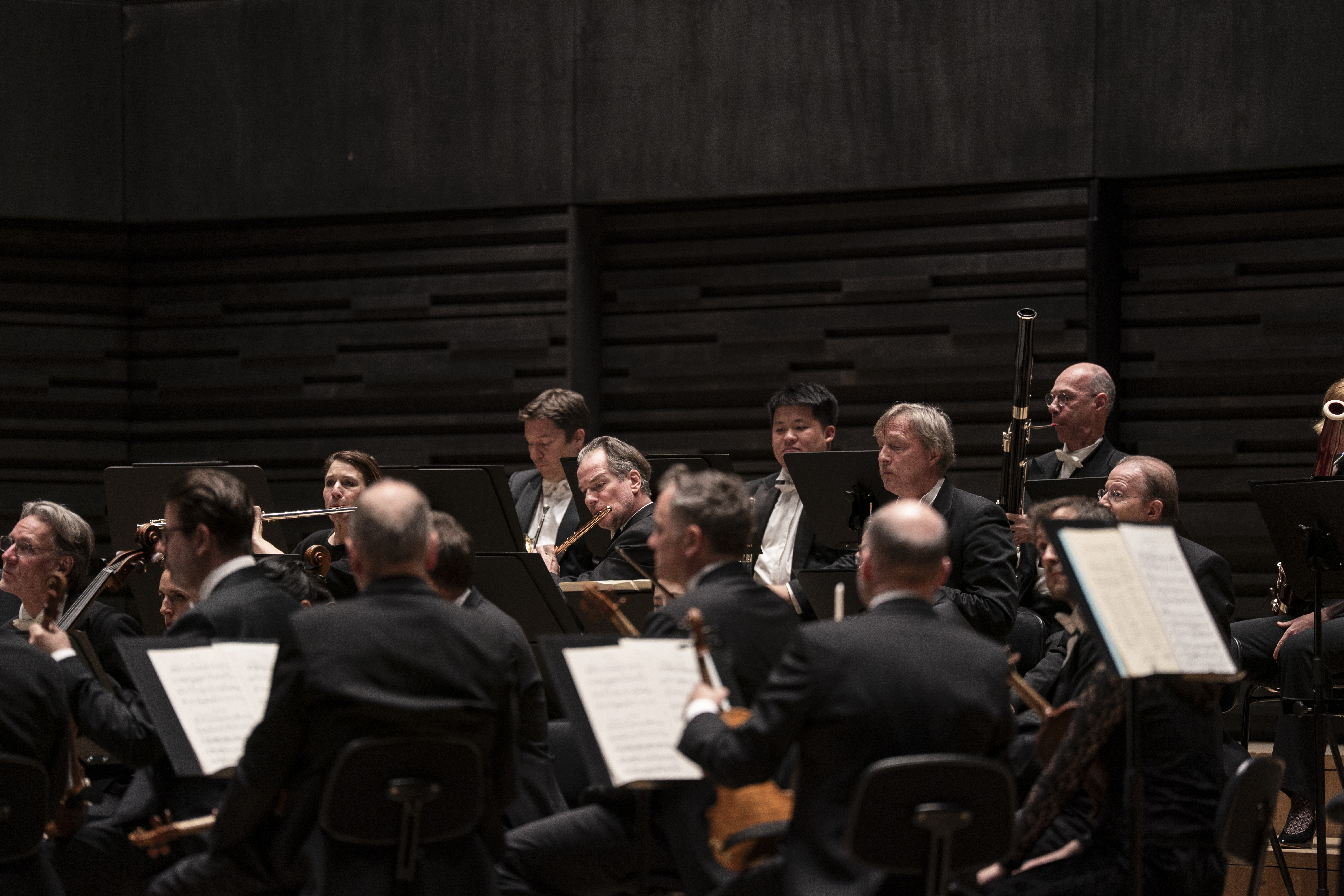 Detailansicht der Holzbläsergruppe eines Orchesters mit Fokus auf Soloflötisten Emmanuel Pahud. Musiker spielen Flöte, Oboe und Fagott. Die Perspektive zeigt die Musiker von der Seite hinter den Notenständern der Streicher in einem schlichten, dunkel getäfelten Saal.