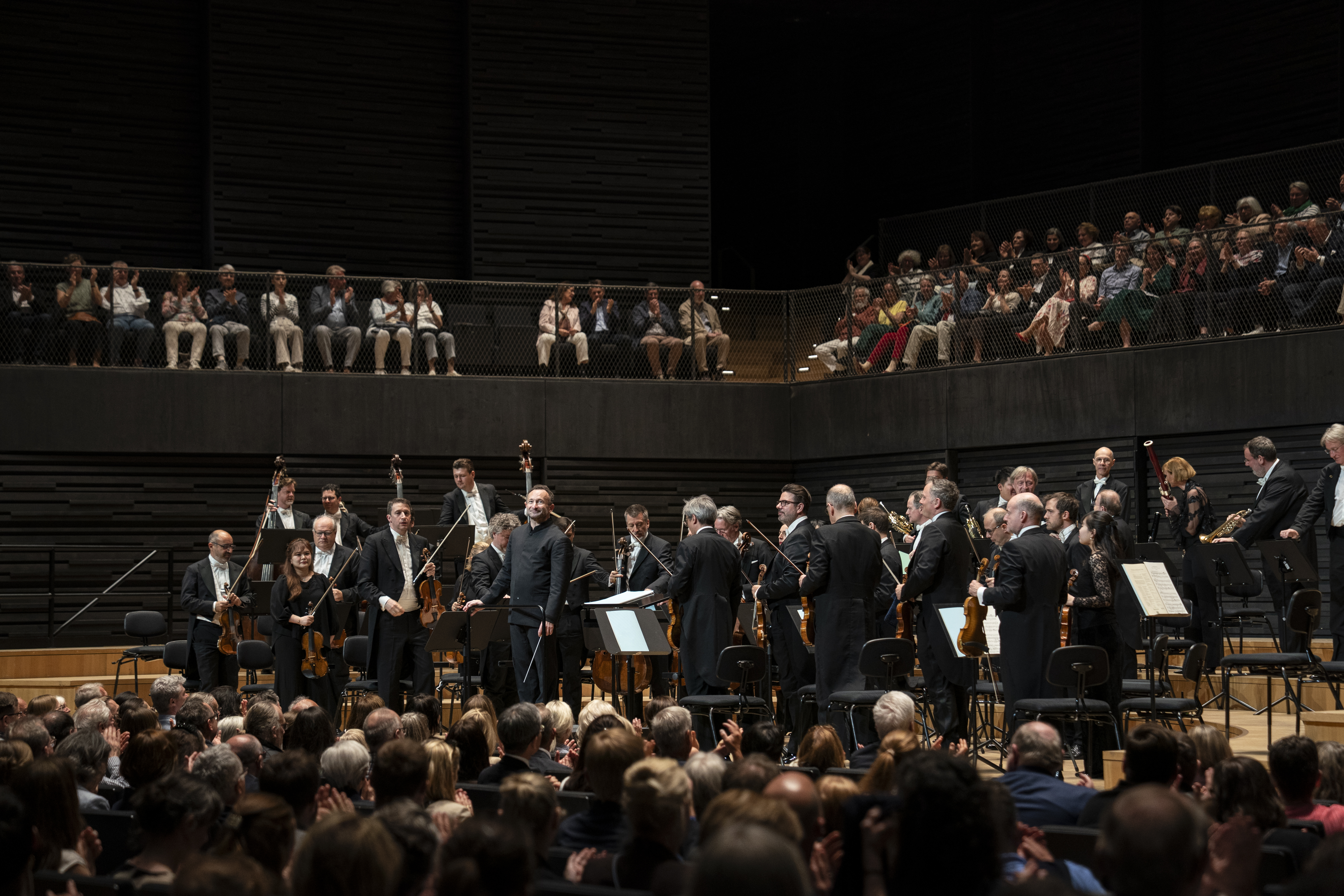 Das Orchester und der Dirigent verbeugen sich nach einem Stück. Das Publikum im Vordergrund und auf den umlaufenden Balkonen applaudiert. Die Architektur ist durch dunkle, horizontale Holzlamellen und eine warme Beleuchtung geprägt.