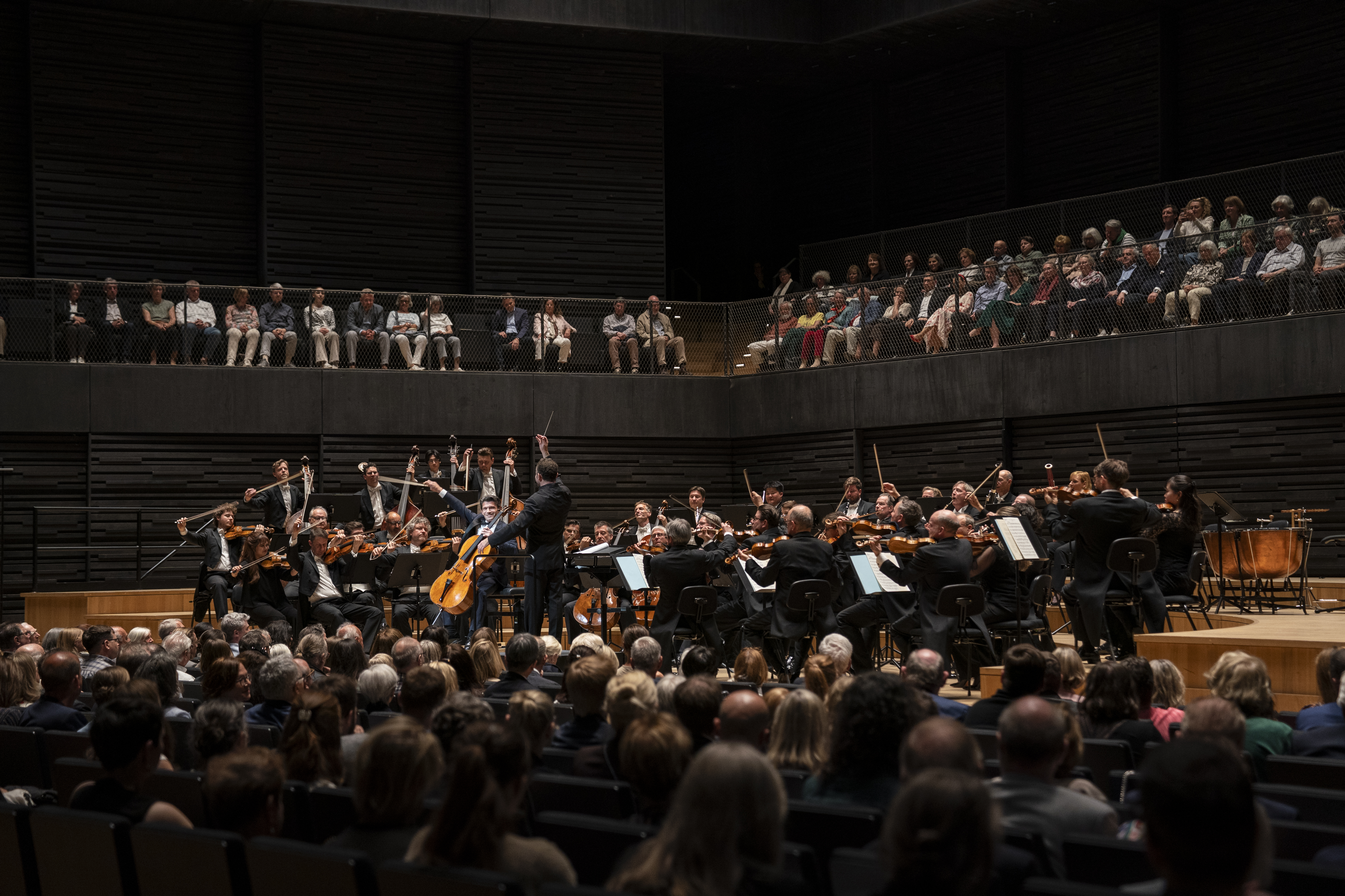 Cellist Gautier Capuçon steht im Rampenlicht neben dem Dirigenten Kirill Petrenko und spielt ein Solo, während das restliche Orchester aufmerksam begleitet. Die Szene ist aus der Mitte des Zuschauerraums aufgenommen und zeigt die fokussierte Atmosphäre während der Aufführung.