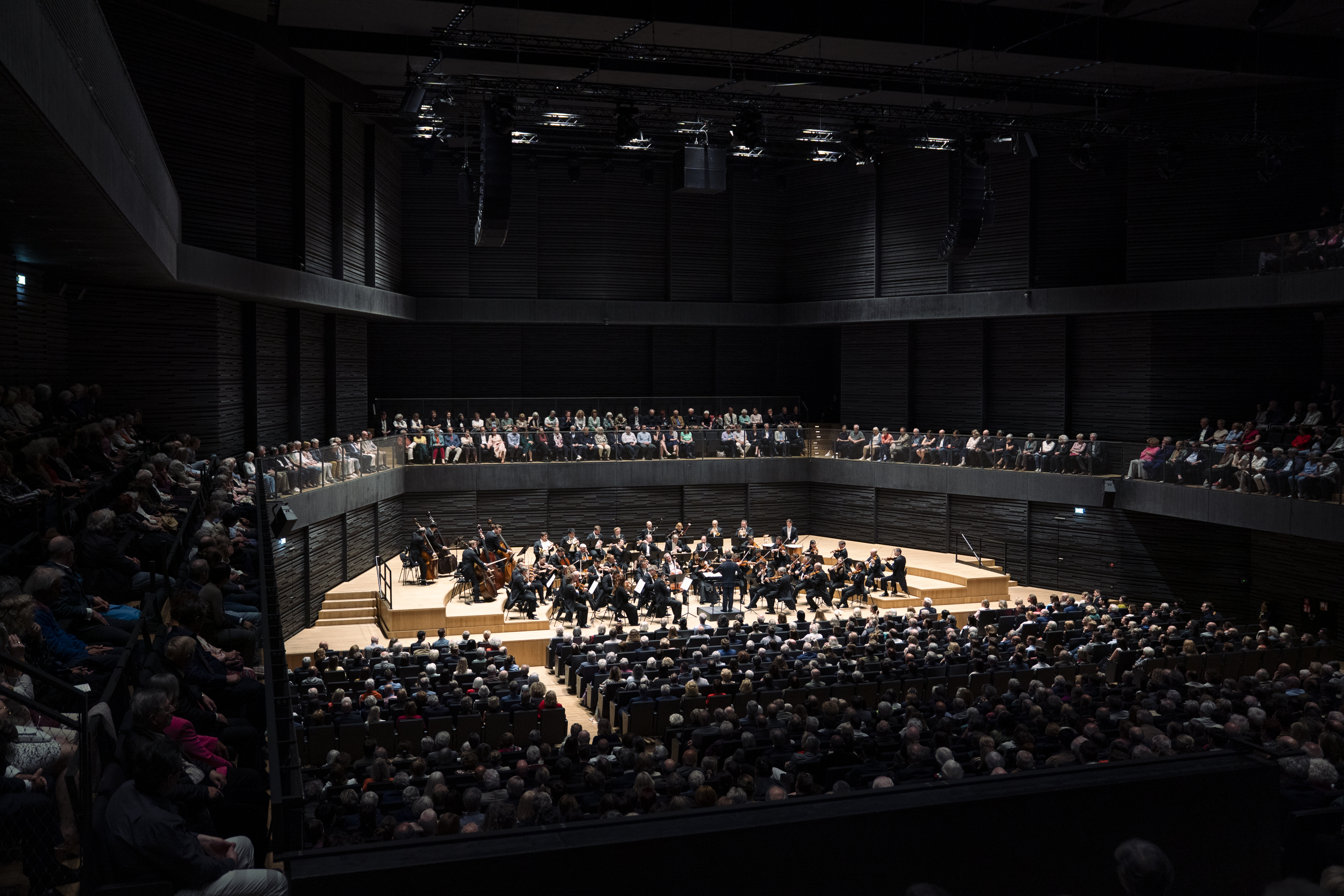 Weitwinkelaufnahme des modern gestalteten, dunklen Konzertsaals. Das Orchester spielt auf einer hellen Holzbühne, umgeben von einem großen Publikum im Parkett und auf den Rängen. Die Beleuchtung konzentriert sich stark auf die Bühne.