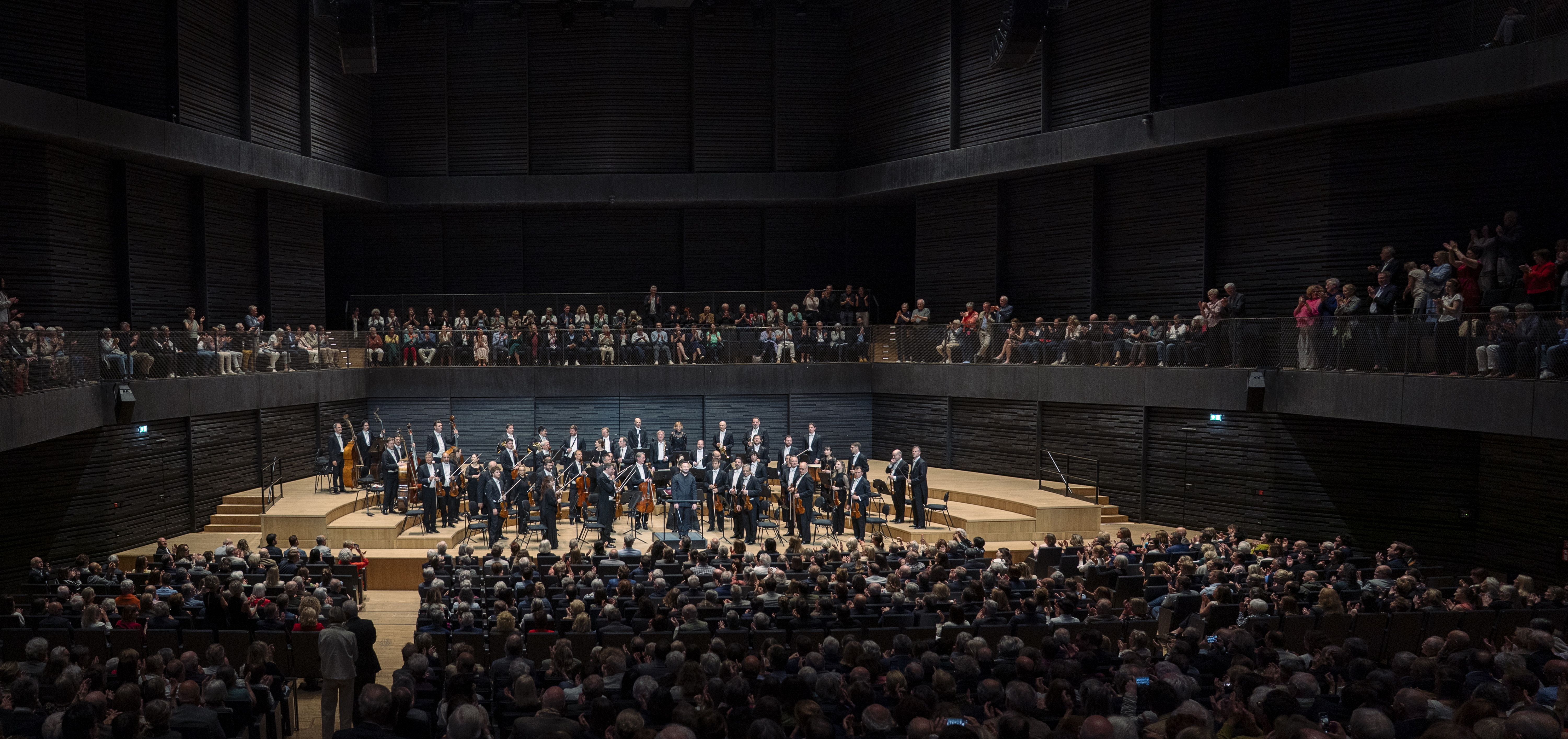 Das gesamte Orchester hat sich auf der Bühne erhoben und nimmt den stehenden Applaus des Publikums entgegen. Die Aufnahme zeigt die imposante Höhe des dunklen Saals und die voll besetzten Zuschauerränge, die die Bühne umschließen.