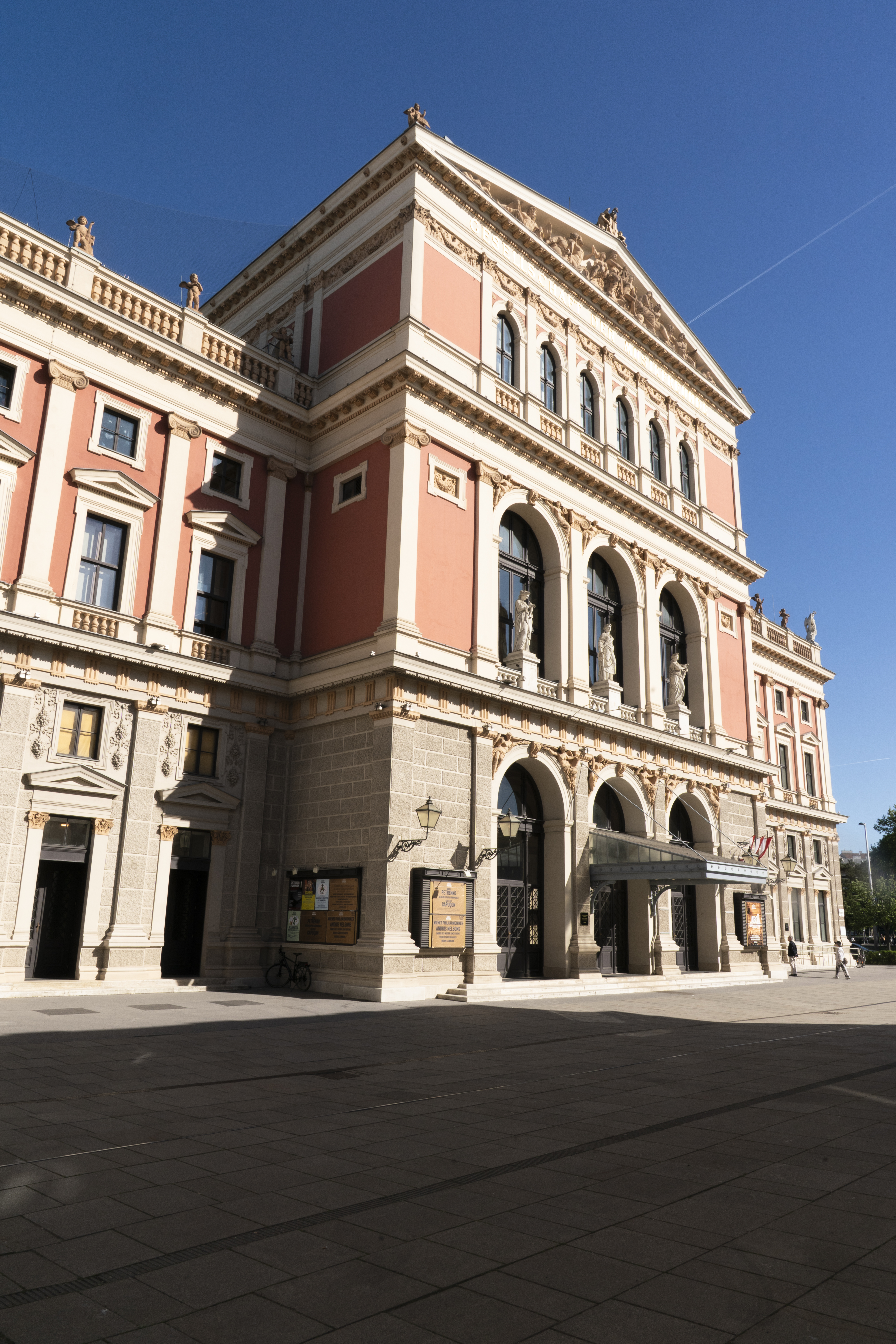 Außenansicht des Wiener Musikvereins bei strahlendem Sonnenschein. Das neoklassizistische Gebäude besticht durch seine lachsfarbene Fassade, helle Steinelemente, korinthische Säulen und Statuen auf dem Dachsims vor einem klaren blauen Himmel.