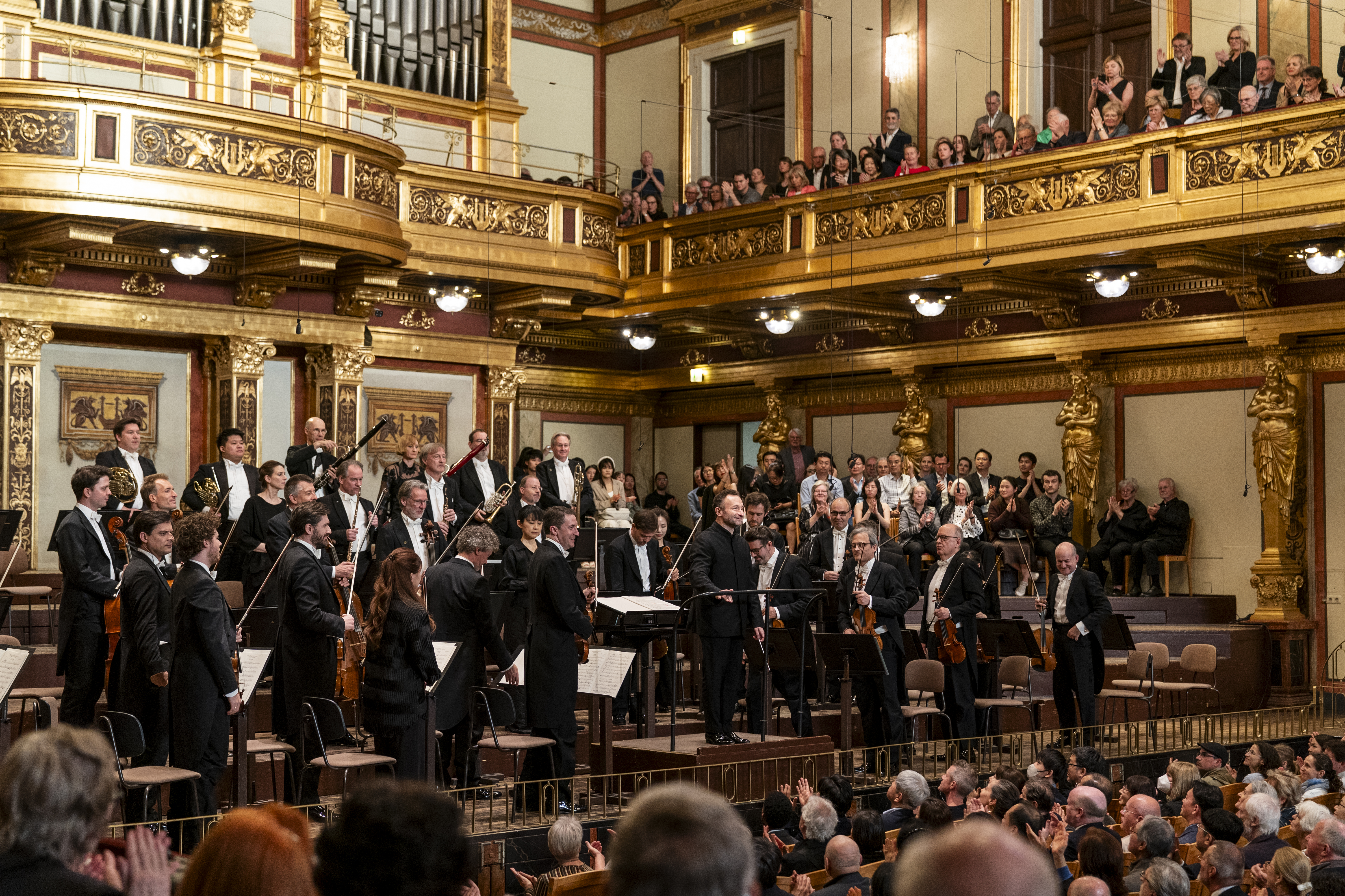Ein Orchester steht auf der Bühne des Goldenen Saals im Wiener Musikverein und verbeugt sich vor dem applaudierenden Publikum. Der Dirigent steht im Zentrum auf einem Podest, umrahmt von Musikern in festlicher Abendgarderobe vor der prachtvollen Orgel.