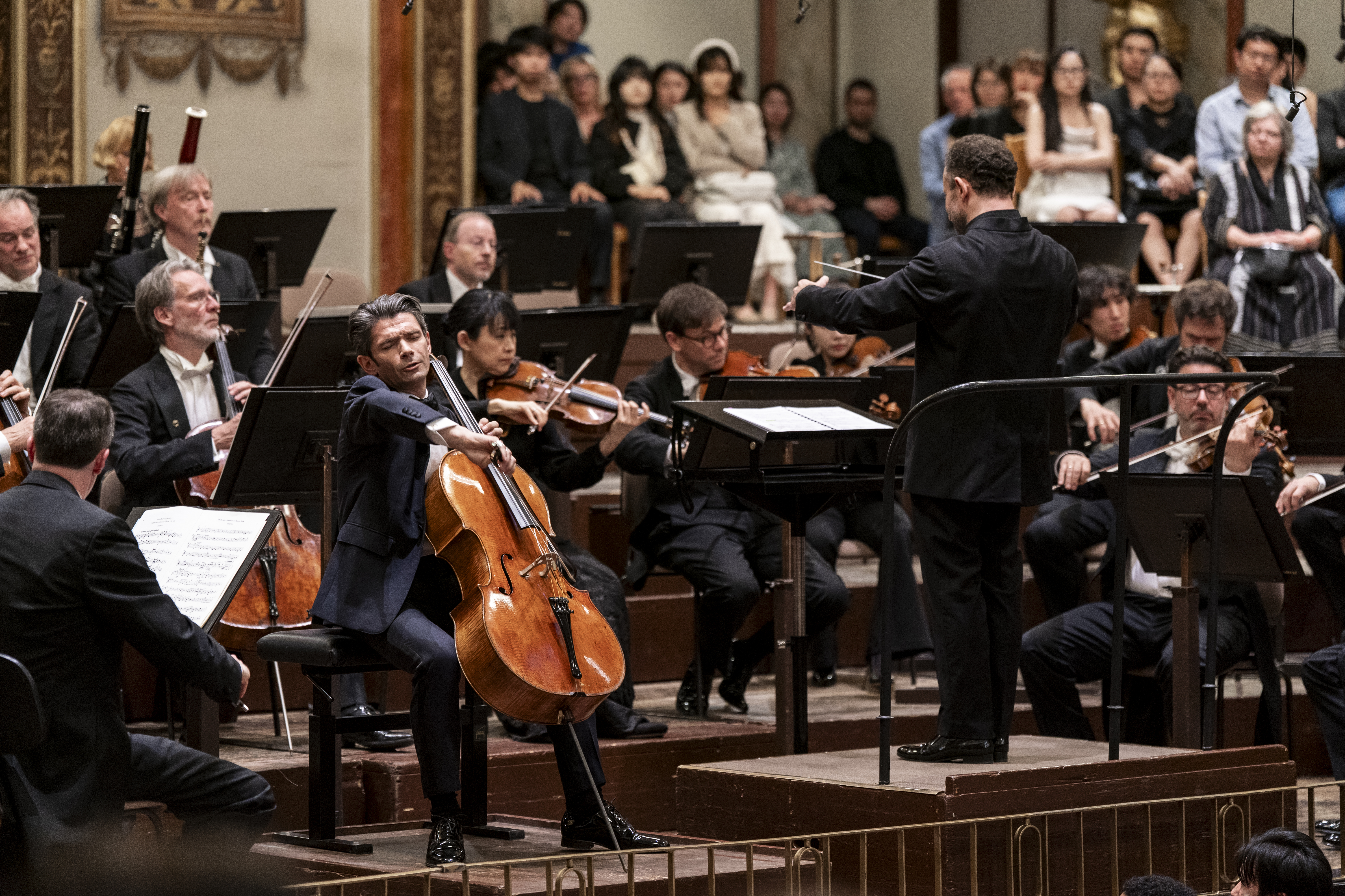 Nahaufnahme des Cellisten Gautier Capuçon während eines Solos oder einer intensiven Passage. Er blickt mit geschlossenen Augen nach oben, während Dirigent Kirill Petrenko rechts im Bild mit dem Rücken zum Betrachter den Takt angibt. Im Hintergrund sind weitere Orchestermitglieder und Zuschauer erkennbar.